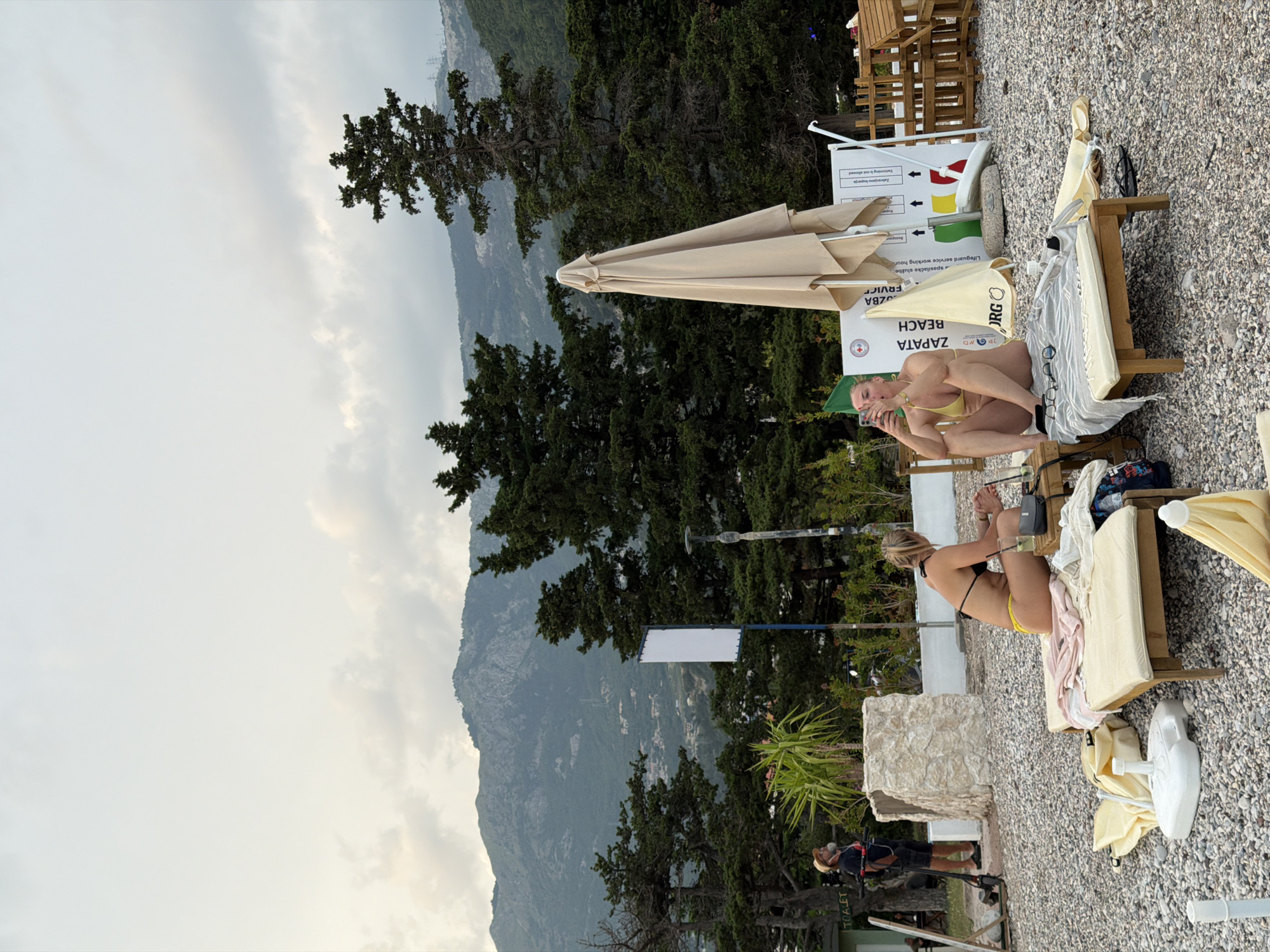 This image depicts a coastal scene on Zapata Beach in Budva, Montenegro. The foreground is dominated by a pebbly beach with two sun loungers. On the left lounger, a woman in a yellow bikini is seated, looking at her phone. On the right lounger, another woman, also in a yellow bikini, is seated, holding a phone. A folded beach umbrella stands behind them, along with a white sign that reads "ZAPATA BEACH". In the background, dense green trees line the base of a mountainous landscape under a cloudy sky. To the far left, a person in a hat stands near a scooter, and further back, a sign reading "TOILET" is partially visible. The overall atmosphere suggests a relaxed day at the beach, with the mountains providing a scenic backdrop.