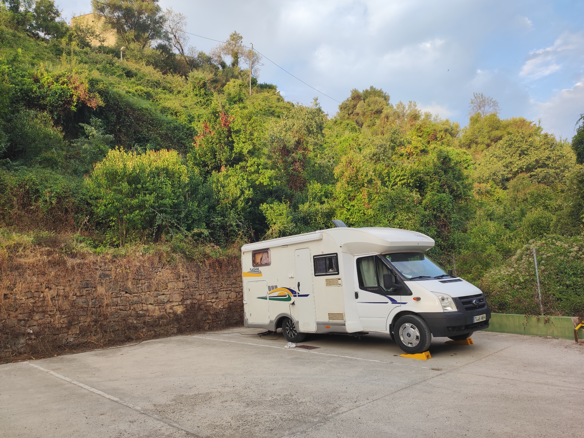 A white camper van is parked in a paved lot at the base of a densely vegetated hillside. The van is positioned facing right, with its front slightly angled towards the viewer. A power cable extends from the side of the van to a point just off-camera to the left. The hillside behind the van is covered in a variety of green foliage, with some patches of dry, brown vegetation and a low stone wall running along its base. A few trees are visible at the top of the hill, and a building or structure can be seen in the upper left corner. The sky is partly cloudy. A pair of white flip-flops are visible on the ground near the van's rear tire. The license plate on the front of the van reads "7744 MBH". The scene appears to be outdoors, likely in a semi-rural or natural setting. The overall impression is one of a temporary stop or campsite.