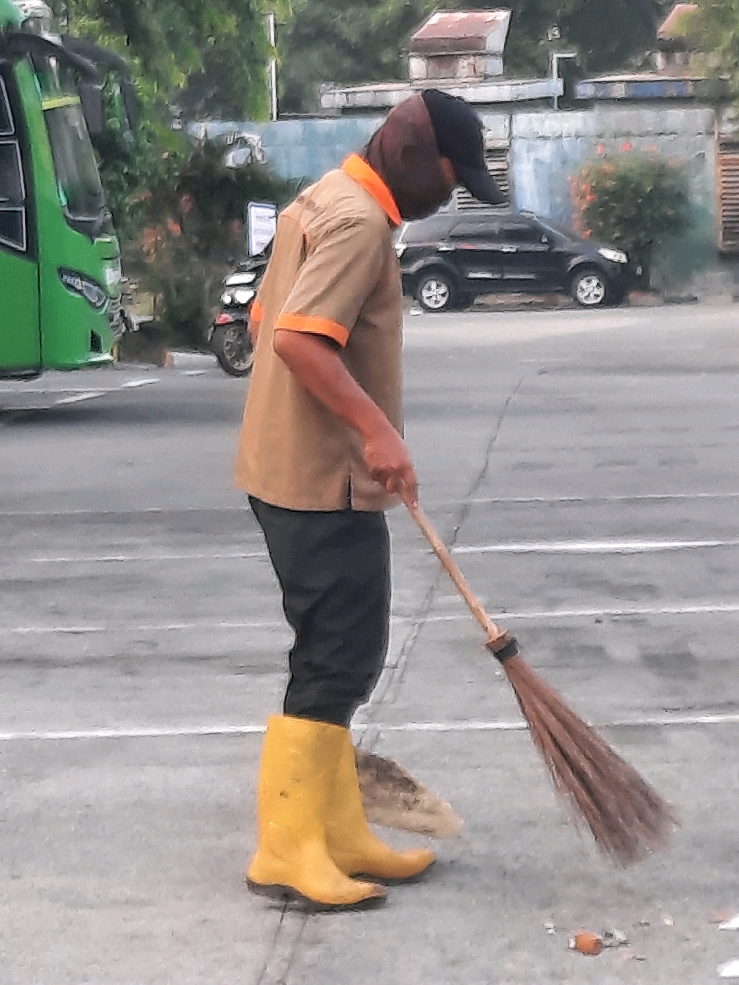A person wearing a brown shirt with orange trim, black pants, a black baseball cap, and yellow rubber boots is sweeping the ground with a broom. They are positioned on a paved surface that appears to be a street or parking lot, with white lines visible on the ground. In the background, a green bus is parked on the left, and a black SUV is visible further back. Various buildings and vegetation are also present in the background, suggesting an urban or semi-urban environment. The scene appears to be captured in East Jakarta, Indonesia.