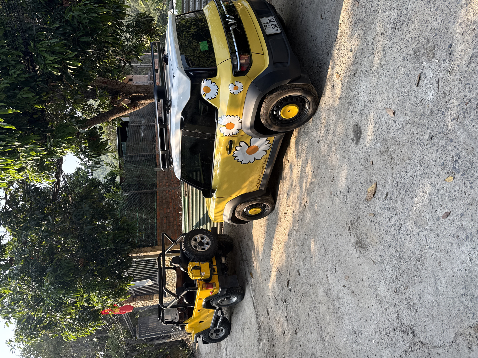 The image shows a yellow vehicle adorned with daisy stickers parked on a concrete surface alongside a smaller, open-top yellow vehicle. A tree with dense foliage stands nearby, and a brick building with a fenced area is visible in the background. A red flag is attached to the smaller yellow vehicle. The license plate on the larger vehicle reads "79A 480.78". The photo was taken in the city of Bac Natural Heritage Area Trang, Vietnam.
