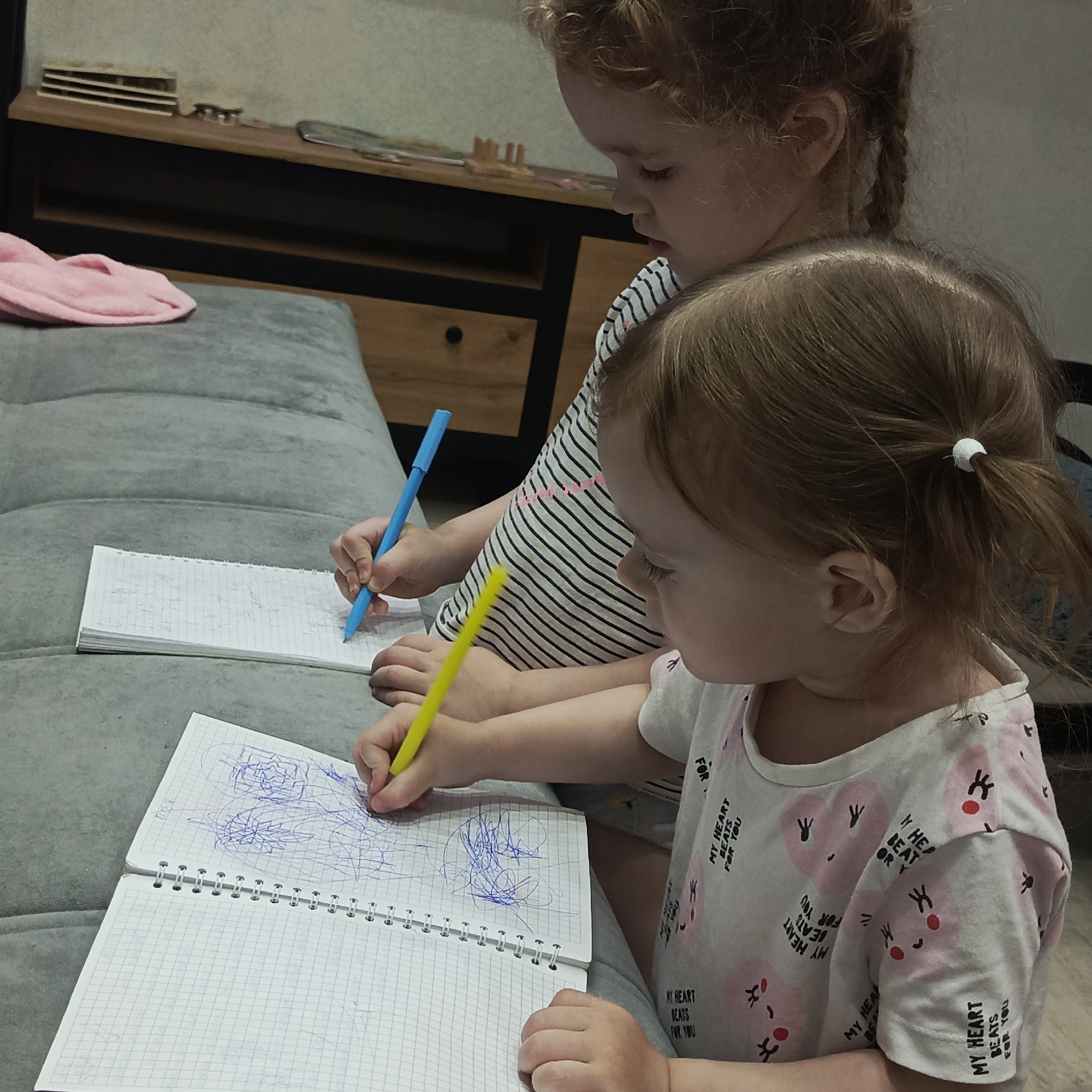 In the city of Марковский, Russia, two young children are depicted seated on a grey tufted upholstered surface, actively drawing in notebooks. The child in the foreground, with light brown hair secured in a small ponytail with a white elastic, wears a white short-sleeved t-shirt adorned with pink hearts and the repeating phrase "MY HEART BEATS FOR YOU." This child is holding a yellow pen in the right hand and is applying blue ink scribbles to the right page of an open, grid-lined spiral notebook. Positioned behind and to the left, the second child, with reddish-brown hair styled in visible braids, wears a black and white striped t-shirt. This child holds a blue pen in the right hand, also drawing in a similar grid-lined spiral notebook. Both children are focused downwards on their respective tasks. To the far left of the grey upholstered surface, a folded pink cloth object is visible. The background reveals a dark wooden low cabinet with a lighter wooden top surface. Several small, light-colored wooden items are arranged on the cabinet top, along with other indistinct objects further to the right. A light-colored wall is visible behind the furniture.