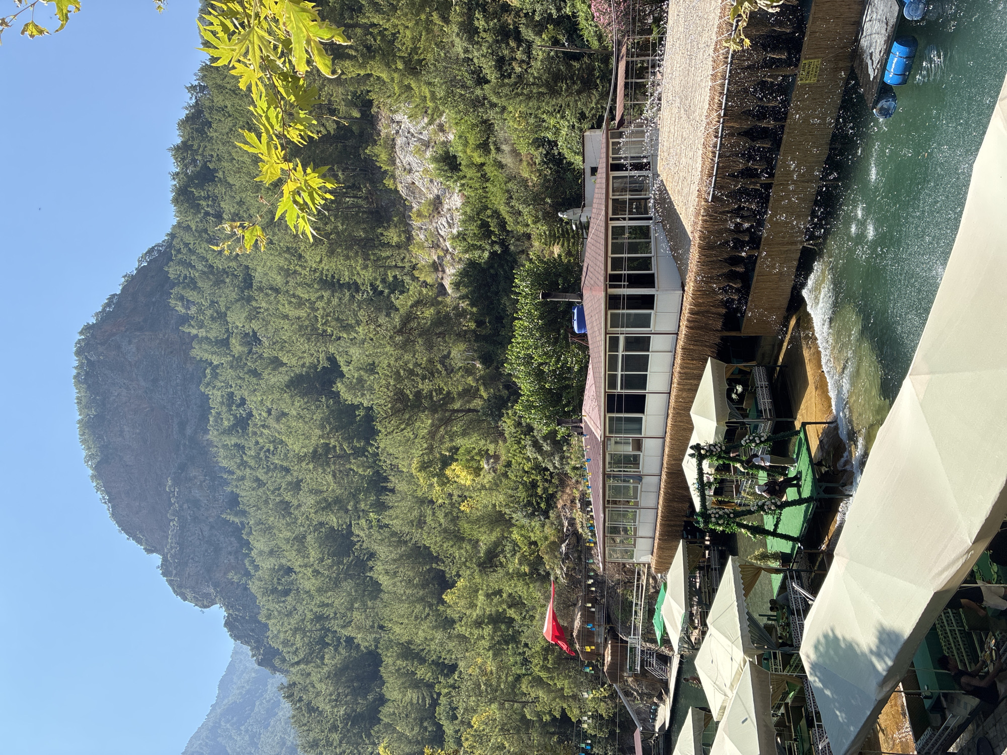 This image depicts a scenic riverside restaurant situated amidst lush green forested mountains under a clear blue sky. The restaurant features a large, modern building with a brown roof, numerous windows, and outdoor seating areas. Several covered dining platforms, some with thatched roofs, are built over the clear turquoise water. These platforms are equipped with seating, tables, and umbrellas, creating a serene dining experience. A swing decorated with greenery and flowers is set up on one of the platforms. The river flows gently, with water cascading down a man-made feature, creating a pleasant ambiance. The surrounding environment is characterized by dense pine forests covering steep mountain slopes, with rocky outcrops visible in the background. The location is identified as Alanya, Turkey, suggesting a natural attraction in the region.