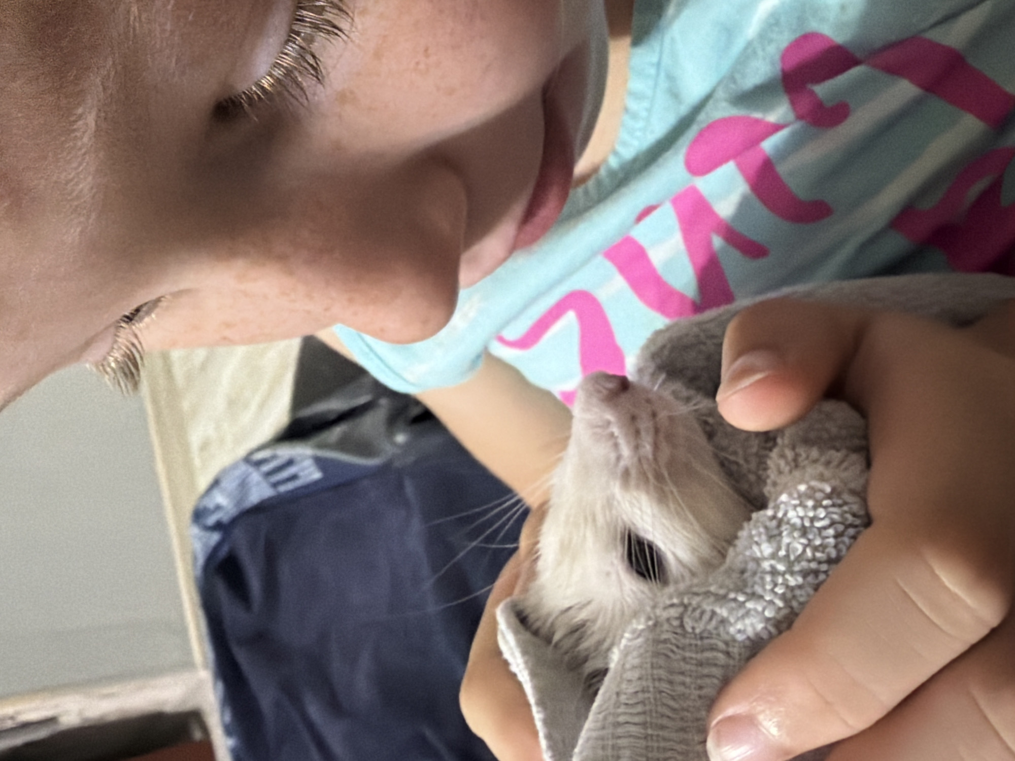 A child with freckles and blonde eyelashes looks down at a small white animal wrapped in a gray towel. The child wears a light blue t-shirt with pink lettering. The animal's face is visible, with dark eyes and whiskers. The background shows a dark blue blanket or fabric. The image is a close-up shot, focusing on the interaction between the child and the animal. The scene appears to be indoors, possibly in a home environment. The location in Dubovaya, Russia is not discernible from the image.