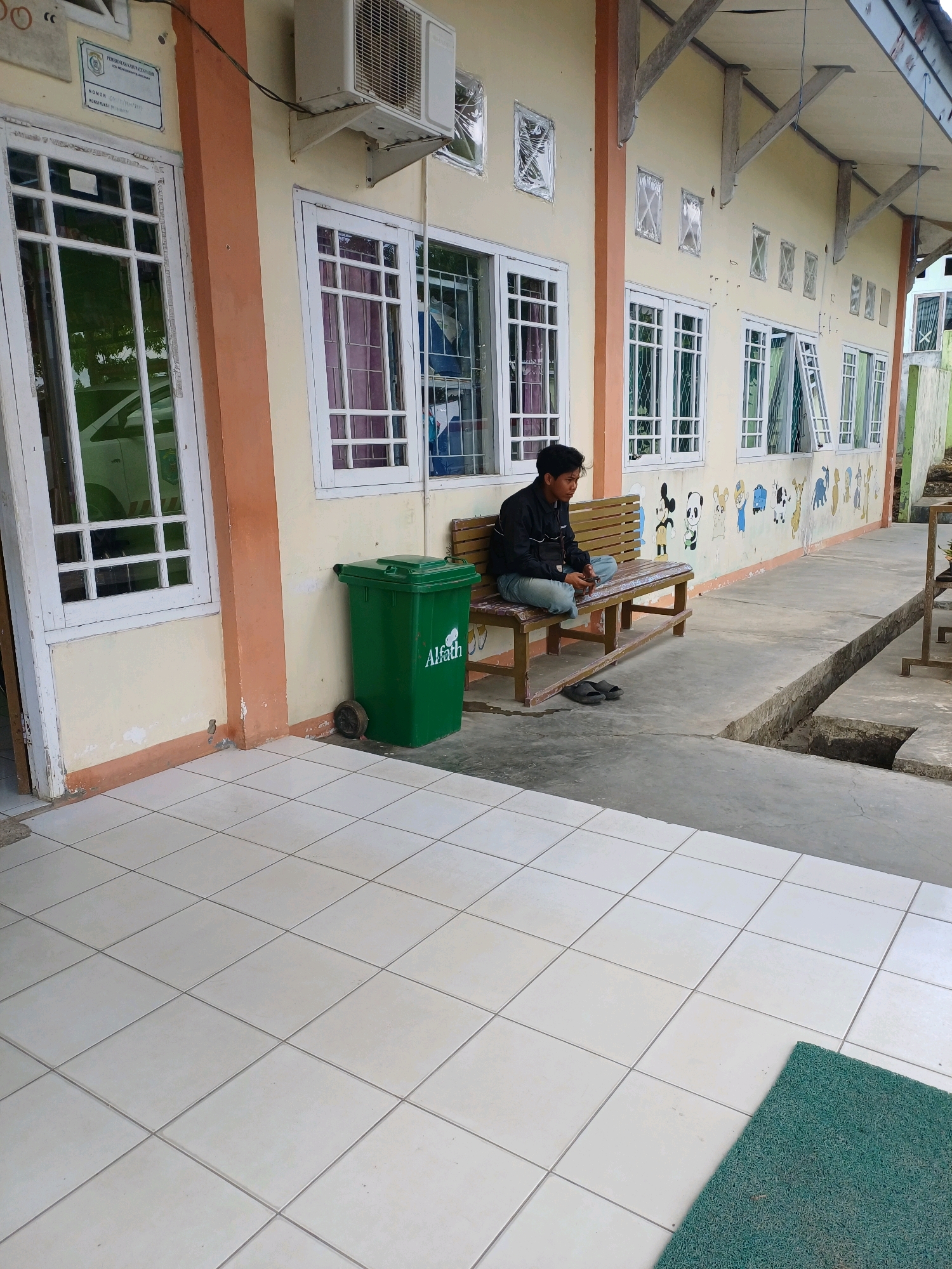 A young man in a black jacket and blue jeans sits on a wooden bench outside a building with a yellow facade. He is looking down at his phone. To his left is a green trash bin with "Alfath" written on it. The building has several windows with white frames and the facade is decorated with cartoon animal drawings. The ground is paved with white tiles. The scene is in Pasir Belengkong, Indonesia.