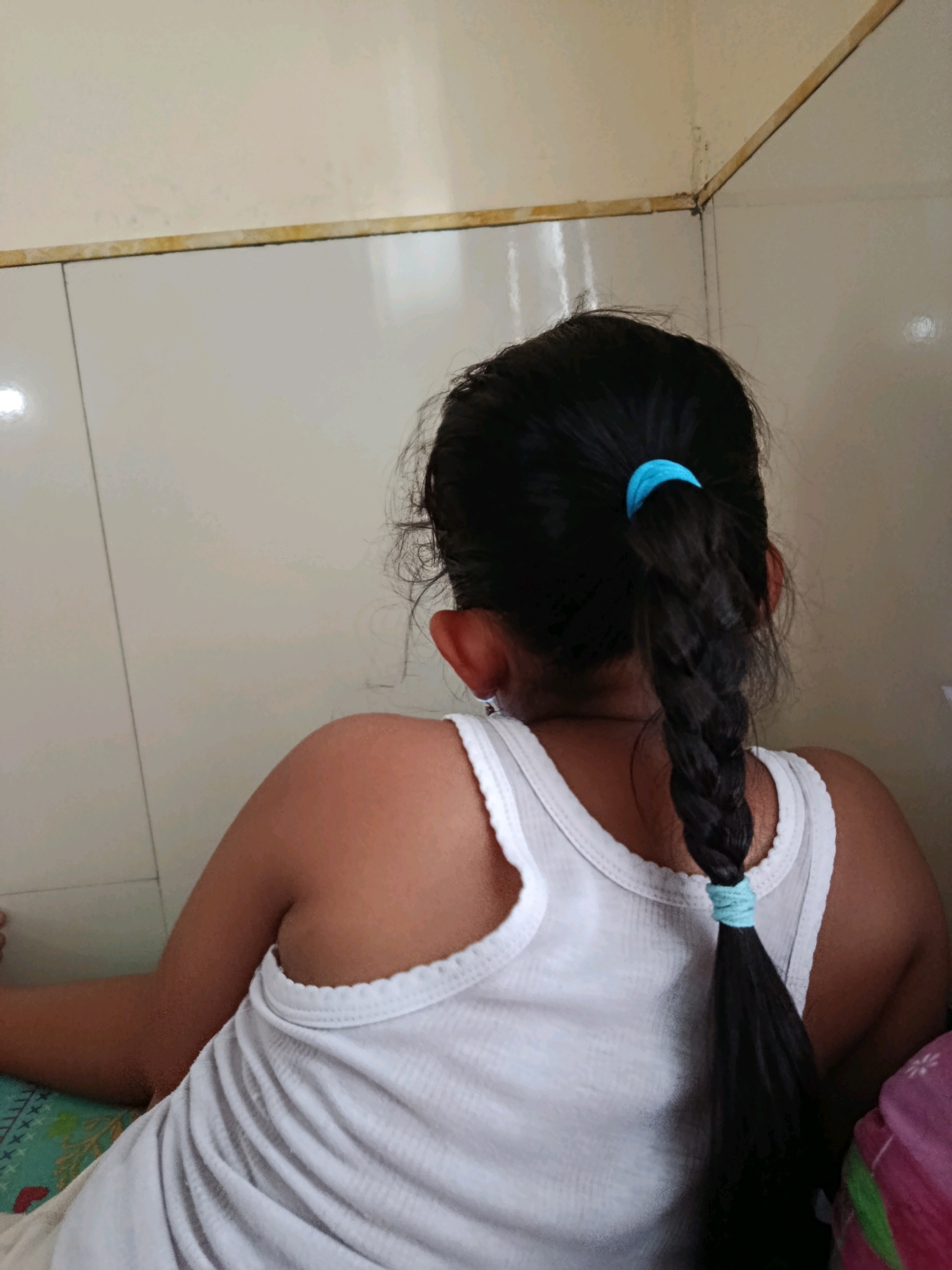 The image shows the back of a young person with dark hair braided and secured with a blue hair tie. They are wearing a white tank top. The background consists of tiled walls, possibly in a bathroom or kitchen, with a wooden trim along the top. The lighting suggests an indoor environment. The scene appears to be located in a home in the city of Karang Rejo, Indonesia, based on the provided location context.