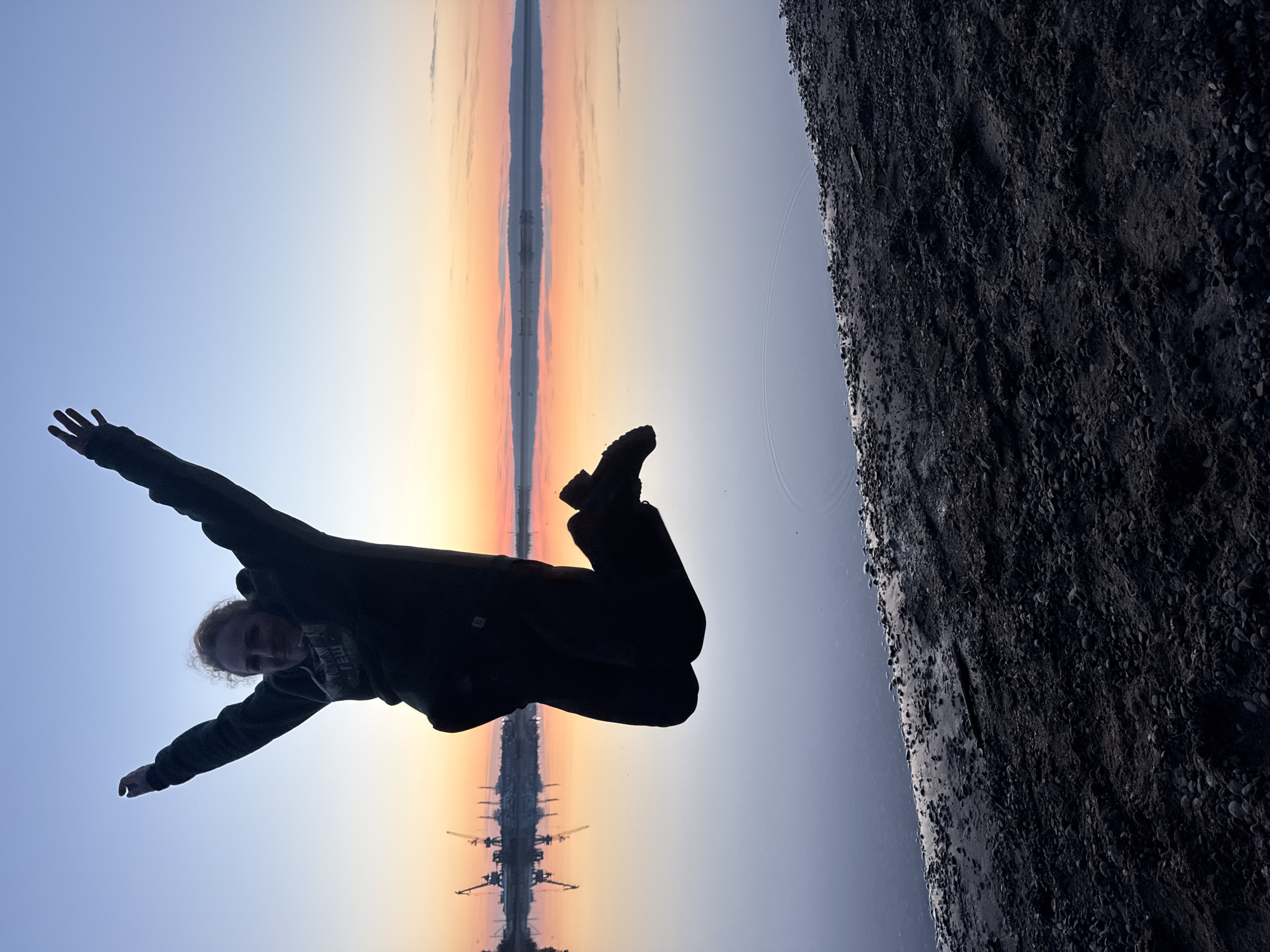 The image captures a dynamic scene with a person silhouetted against a vibrant twilight sky, positioned as if flying or falling, with their arms outstretched and one leg bent. The photo is oriented vertically, with what would typically be the sky on the left and the ground on the right, creating a unique perspective.
The central subject is a person, likely a young adult, with light-colored hair, wearing dark clothing. Their form is mostly a dark silhouette against the bright sky, but their face is slightly visible, looking towards the camera with a subtle expression. Their arms are spread wide, and their right leg is lifted and bent, suggesting a joyful jump or a playful pose in mid-air.
The background features a stunning gradient sky, transitioning from a soft blue at the top-left to warm orange, pink, and yellow hues towards the center and right. This color palette indicates either sunrise or sunset. A distinct, narrow structure, possibly a pier, bridge, or industrial line, extends into the distance through the center of the image, creating a strong symmetrical reflection in the calm body of water beneath it. Several cranes are dimly visible along this distant structure. The body of water also reflects the colorful sky.
To the right side of the image, occupying a significant portion, is a dark, textured area of ground composed of small stones or gravel, indicating a rocky shore or beach. The unusual angle enhances the sense of motion and freedom. Given the context, this scenic view is located in Chaikovsky, Russia, possibly along the Kama River or a nearby reservoir, with industrial elements in the distance. No visible text is present in the image.