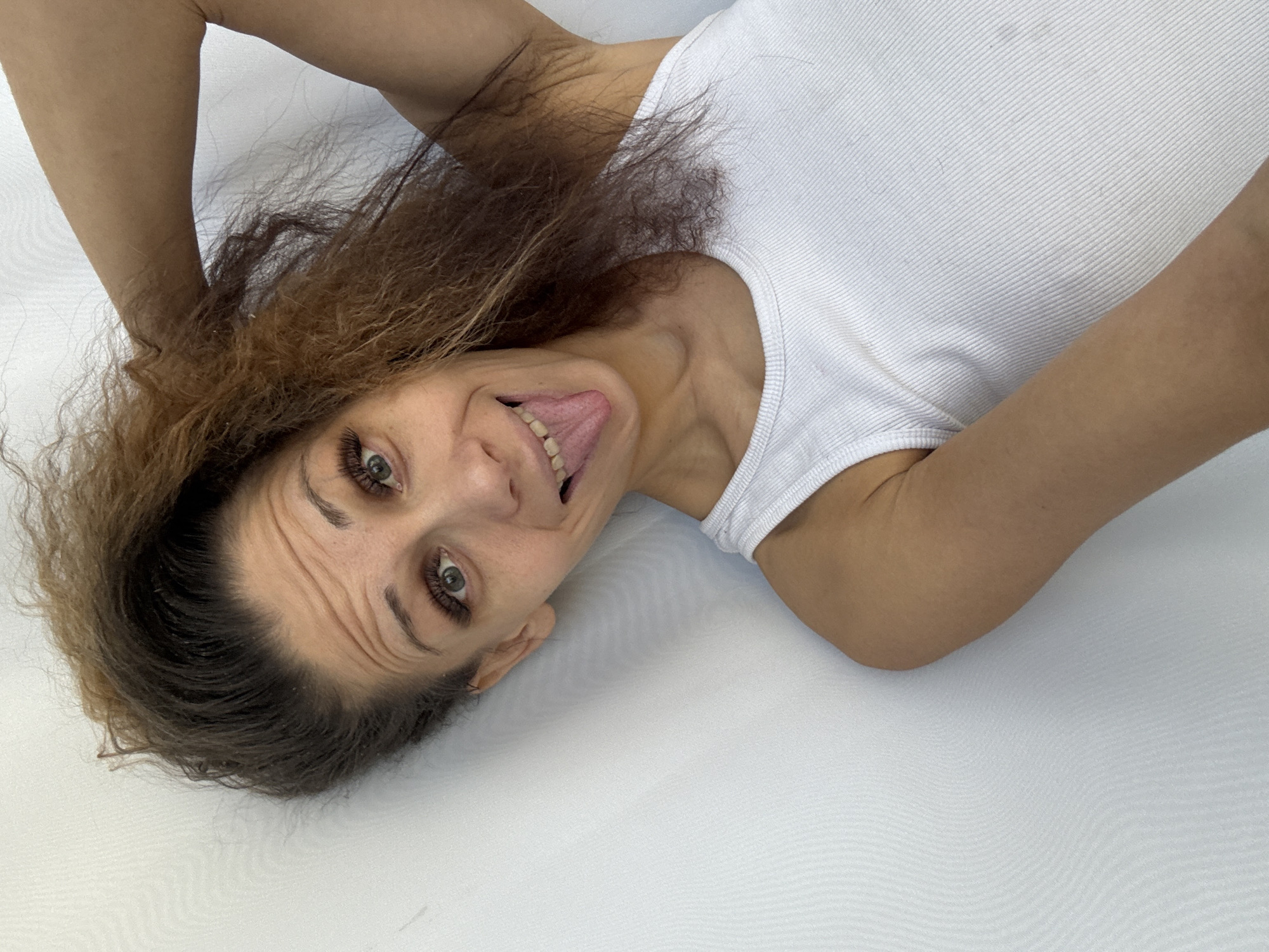 An overhead, close-up shot captures a person, likely a woman, lying on a plain white surface, possibly a bed or mattress. Her head is tilted to the viewer's right, and she is looking directly into the camera with a playful and expressive facial gesture. She has light-colored eyes with prominent eyelashes, and brown, wavy hair that appears somewhat disheveled and spreads around her head. Her mouth is open in a wide smile, revealing her teeth, and her tongue is extended slightly, contributing to her humorous demeanor. Wrinkles are visible on her forehead, enhancing the animated quality of her expression.
The individual is wearing a white, ribbed tank top or camisole. Her arms are partially visible, raised above her head, suggesting a relaxed or spontaneous pose. The background is uniformly white, with a subtle textured pattern visible on the surface she is resting on and extending into the background. The lighting is bright and even, indicating an indoor setting. The overall mood of the image is cheerful, lighthearted, and candid. There is no visible text in the image. The image was provided with the location context of Rusanovo, Russia.