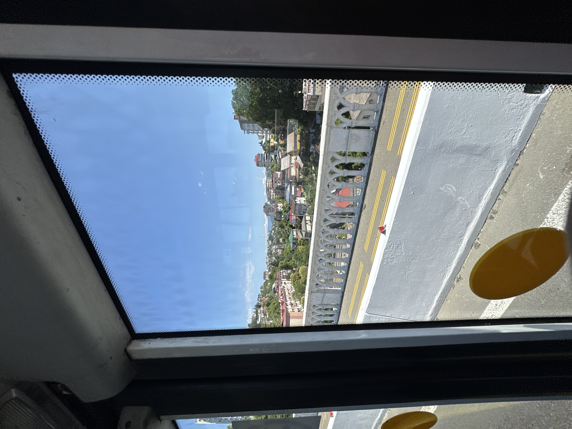 The image shows a view from inside a vehicle, likely a train or bus, looking out a window. The window overlooks a cityscape with buildings on a hillside under a clear blue sky. In the foreground, there is a white railing with an ornate, arched design, characteristic of a bridge or elevated walkway. Yellow lines mark a road or pathway beyond the railing. Two yellow circular stickers are visible on the interior of the window. The architecture of the buildings suggests a location in a warm climate, with red and orange tile roofs common. The general ambiance is that of a bright, sunny day in a coastal or hilly urban area. This scene is consistent with views found in Sochi, Russia, known for its hilly terrain and coastal architecture.