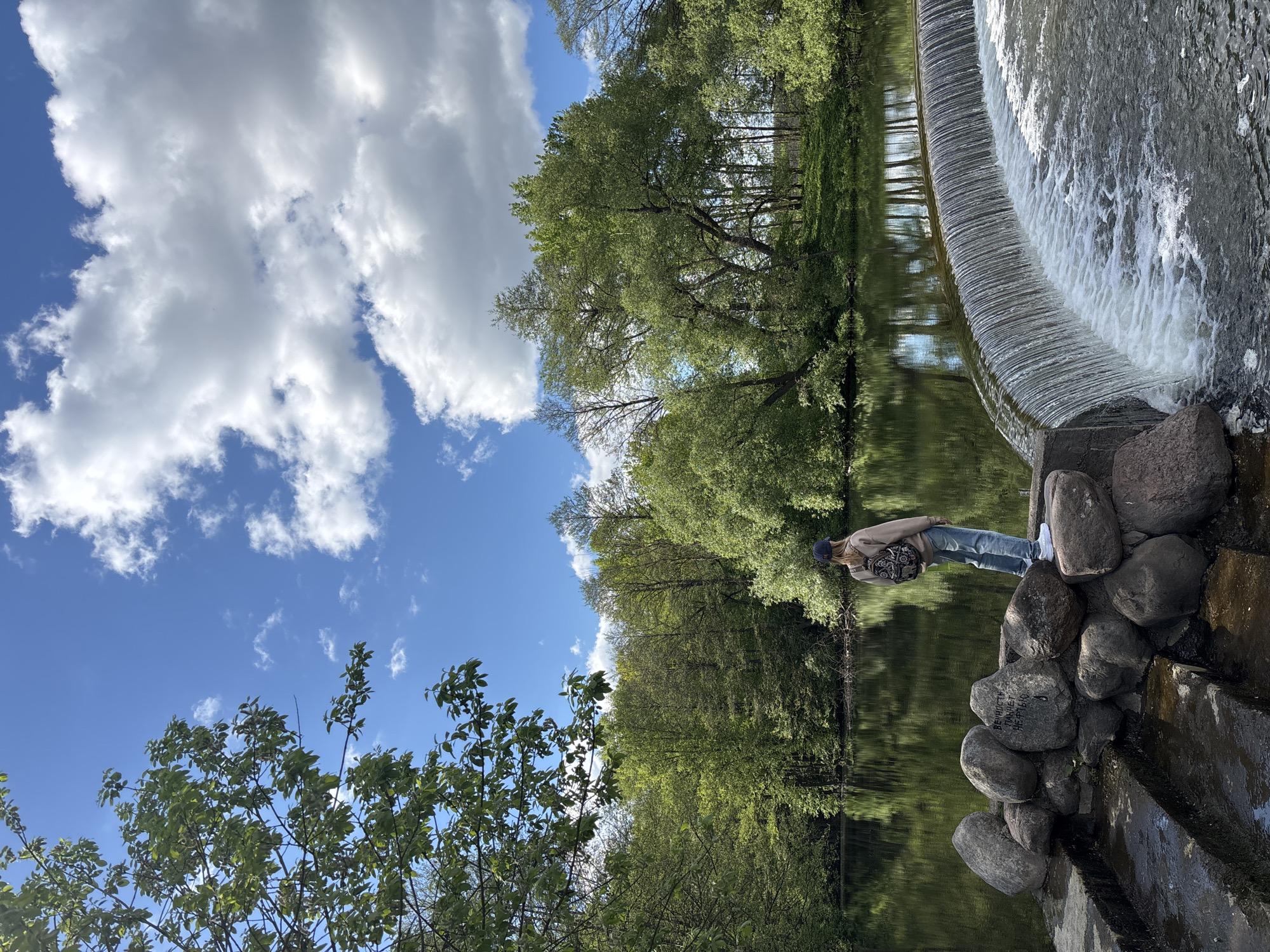 A person stands on a rock formation at the edge of a small dam waterfall. The dam has a curved design and the water falls in a smooth sheet. The person is wearing jeans, a jacket, and a dark cap. The person has a dark backpack, and appears to be looking at the water. The sky is blue with white clouds and trees line the opposite side of the water. The location is Папярнянскі сельскі Савет, Belarus.