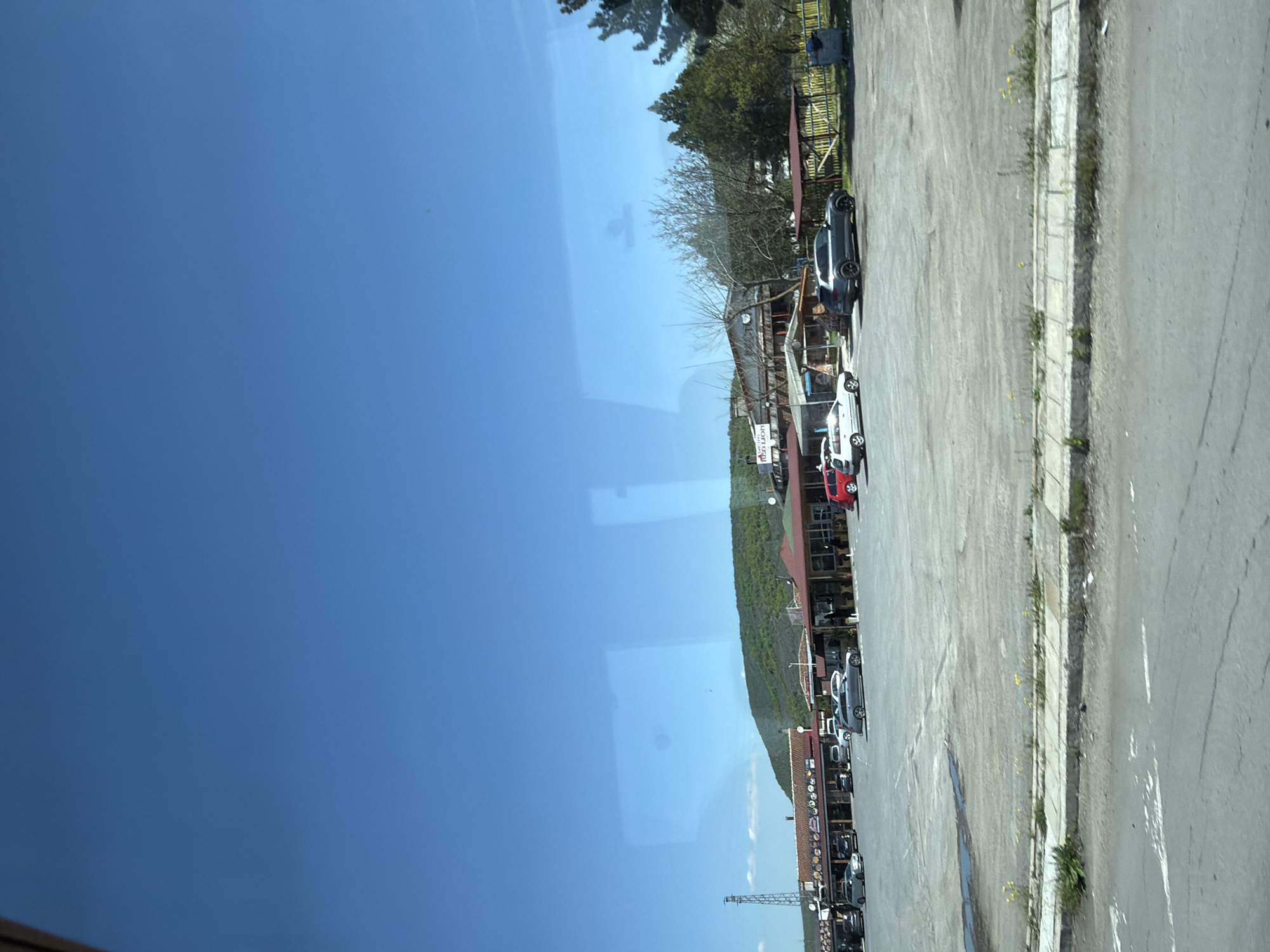 The image shows a street view, possibly taken from inside a vehicle, in Gorno Aleksandrovo, Bulgaria. The sky is clear and blue. A row of buildings lines one side of the street, with cars parked along the roadside. The buildings appear to be commercial establishments, possibly restaurants or shops, judging from the signage and awnings. The road is paved and appears to be moderately used. Vegetation, including trees and shrubs, is visible near the buildings and along the roadside. The scene seems to capture a regular day in the city, with no specific interactions or actions prominently visible. The environment is typical of a small town or village.