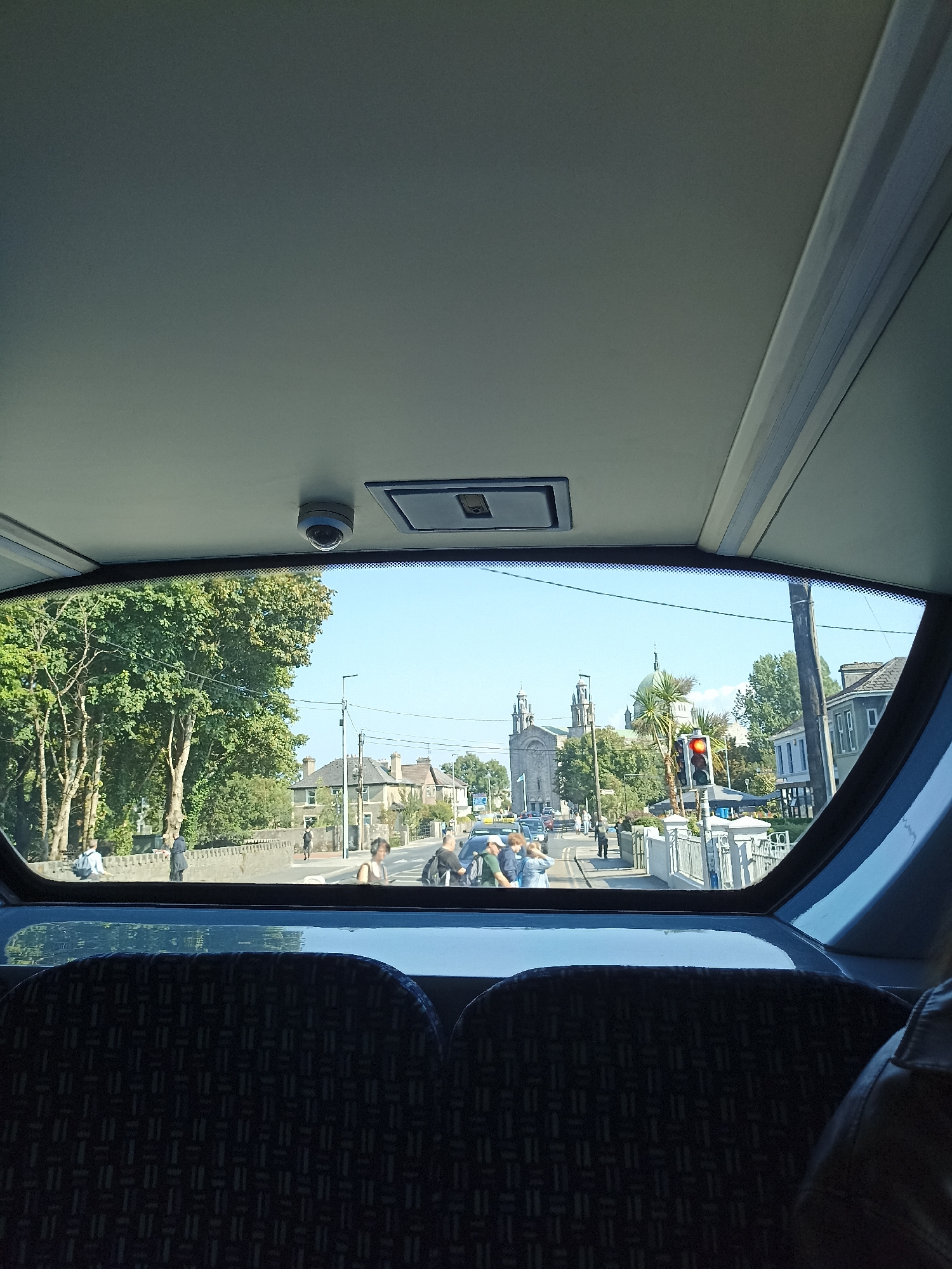 The image captures a view from inside a vehicle, looking out through the rear window. The foreground is dominated by the patterned upholstery of two seats, one on the left and one on the right, suggesting the perspective is from the back of a bus or train.
Through the window, a sunlit street scene unfolds. On the left, lush green trees line a low stone wall. Beyond the wall, several individuals are visible, some walking and others sitting. The middle ground features a street with a traffic light showing red. A large, prominent building with twin spires and a dome, characteristic of a cathedral, is visible in the distance. This architectural style, coupled with the presence of palm trees, strongly suggests the location is the Galway Cathedral in Galway City, Ireland.
Further down the street, more buildings and people are seen, along with cars. The sky is a clear, bright blue. The overall impression is of a bright, pleasant day in an urban environment. The lighting indicates it is daytime, likely late morning or early afternoon given the angle of the sun.