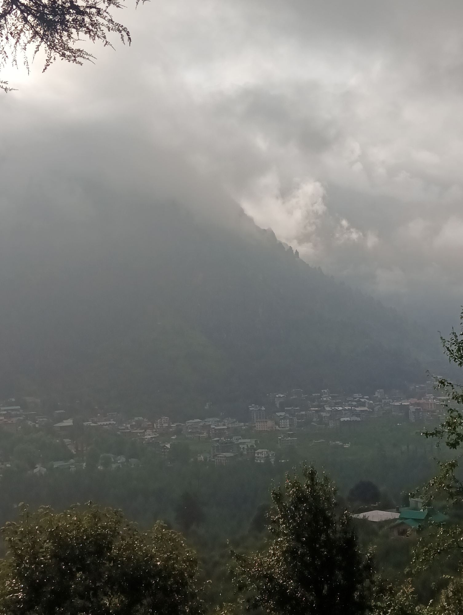The image captures a scenic view of a mountainous landscape in Chhyal, India, with a town nestled in the valley below. The sky is overcast with thick, grey clouds, with a hint of sunlight breaking through the upper left corner. A tree branch with dark leaves is visible in the top left, silhouetted against the bright sky.
The mid-ground features a densely populated town with numerous buildings of varying sizes and architectural styles, including some taller structures and smaller residential houses. The buildings are tightly clustered together, indicating a well-established urban area. The town is surrounded by lush green hills and dense vegetation, suggesting a natural environment.
In the foreground, the image is partially obscured by the dark foliage of trees, which appear to be in the process of being illuminated by the faint sunlight. The overall impression is one of a quiet, possibly misty or rainy day in a beautiful, mountainous region. The scene conveys a sense of depth and scale, with the vastness of the mountains contrasting with the human settlement in the valley.
