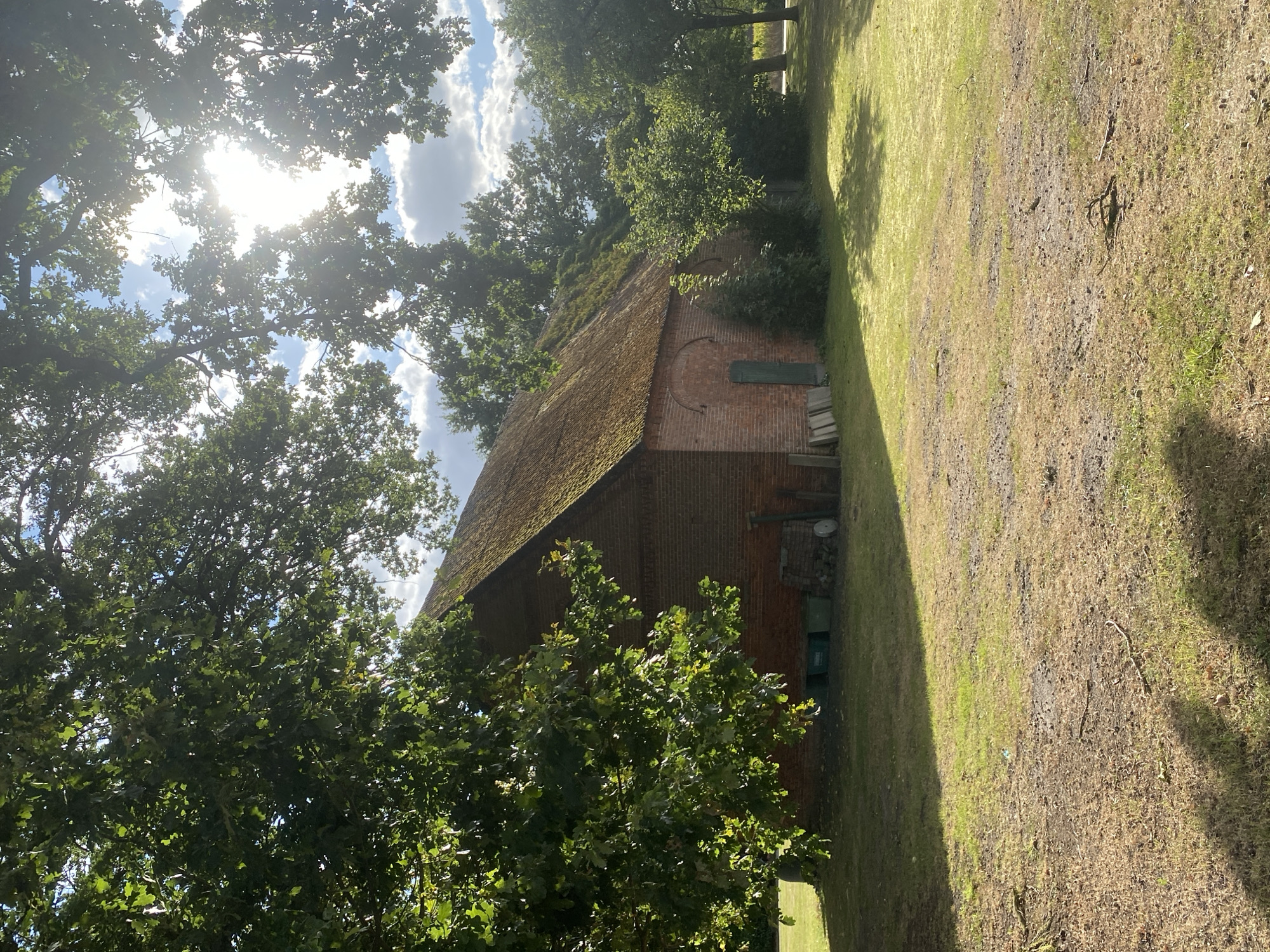 An exterior, eye-level, medium shot captures a weathered brick barn with a moss-covered, pitched roof under a bright, sunlit sky in Schwarme, Germany. The barn's facade is predominantly brick, with a single green door and several arched windows visible. To the left of the barn, a tree with lush green foliage casts a shadow on the dry, patchy grass in the foreground. Various objects, including green bins and what appear to be wooden planks, are placed near the barn. The background consists of dense green trees and a partly cloudy sky, with the sun's rays filtering through the branches. The scene suggests a rural setting, likely a farm or rural property.