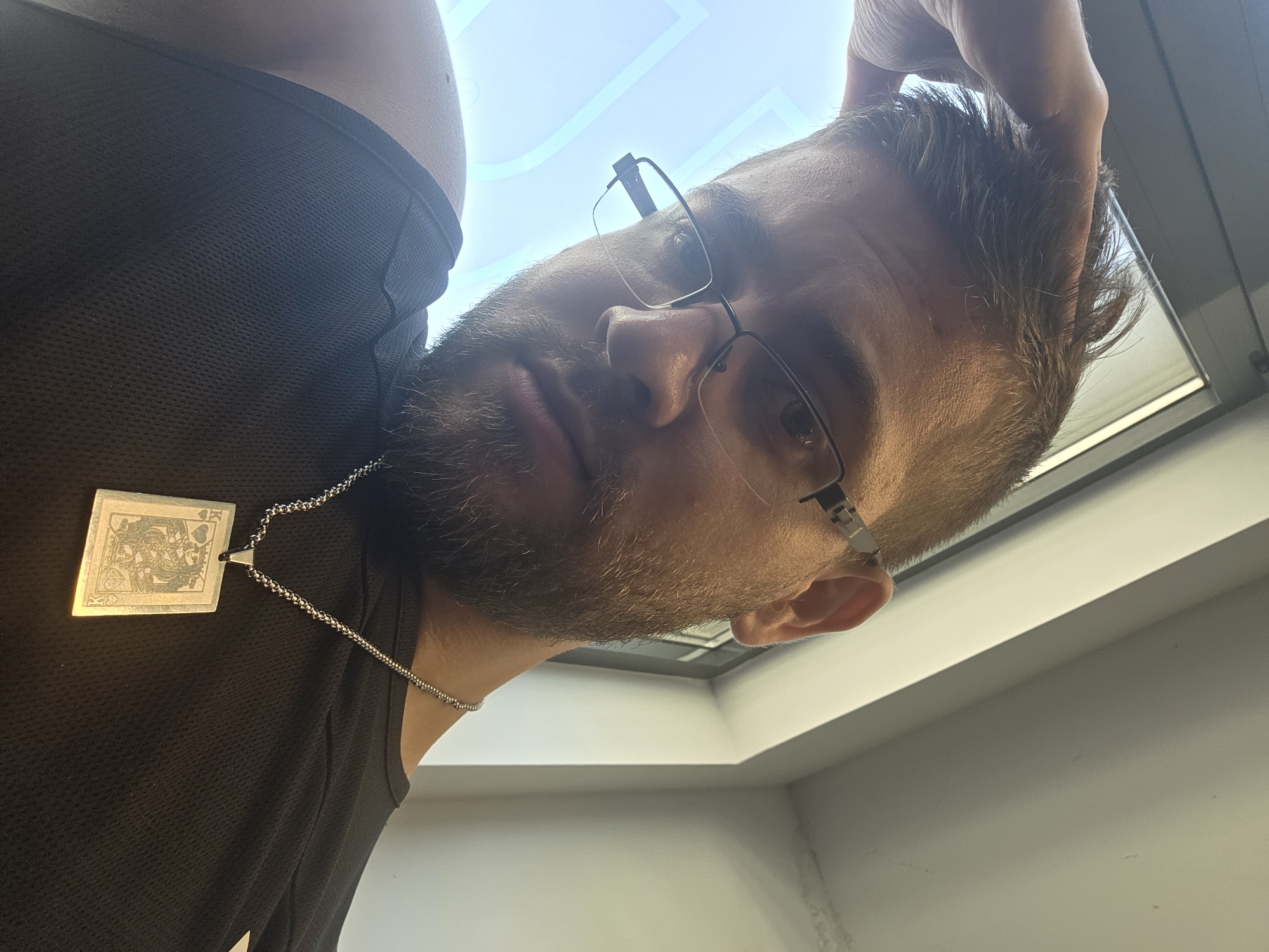The image presents a low-angle perspective of a man, likely taken as a selfie, looking downwards towards the camera. He has a beard and short, brownish hair, and wears rectangular, dark-framed glasses. He is dressed in a dark, textured sleeveless top, which appears to have a mesh-like pattern. Around his neck, he wears a silver-colored chain necklace with a prominent rectangular pendant. The pendant features an etched, metallic design resembling a playing card, specifically a stylized King of Hearts, complete with a regal figure and heart suit symbols. One of the man's hands is visible near his head, seemingly resting on or adjusting his hair.
The setting is indoors, brightly illuminated by what appears to be a large overhead skylight or window, identifiable by the bright light and a subtle geometric pattern on the ceiling. The walls are light-colored, possibly white or light gray. The bright lighting suggests it is daytime. The man's expression is neutral and direct as he gazes towards the camera. The image offers no discernible text on his clothing or in the background, only the symbolic design on the pendant. The context specifies the location as Plovdiv, Bulgaria.