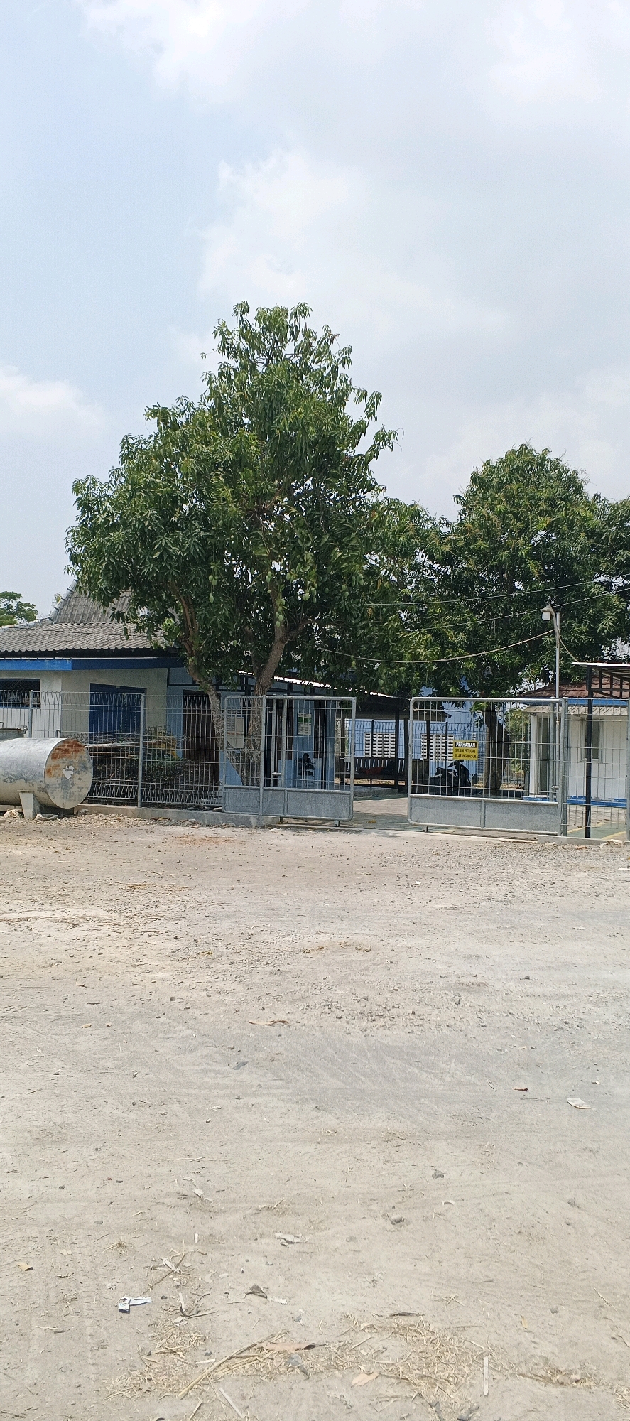 The image depicts the entrance to a facility in Parengan, Indonesia. A large, mature tree with green leaves and some visible fruit stands prominently in front of the gate, partially obscuring a building behind it. The entrance is a large, metal gate, open, allowing access to the facility. The ground in front of the gate is unpaved, with a sandy or dirt-like texture, showing some tire tracks and scattered debris. To the left of the gate, a large, cylindrical metal object, possibly a tank, rests on the ground. The building behind the gate appears to be a single-story structure with a blue trim and a grey roof. The sky above is cloudy. A yellow sign with text is visible on the fence inside the gate.