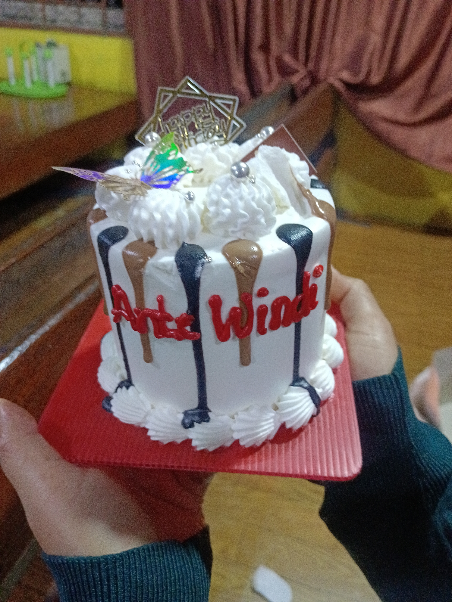 A close-up, eye-level shot shows a person holding a white cake on a red cake board. The cake is decorated with white frosting piping around the bottom edge and around the top, with dollops of frosting on top. Dripping down the sides of the cake are streaks of dark chocolate and milk chocolate frosting. Red lettering on the side of the cake spells out "Arte Windi". A gold "Happy Birthday" topper is on the top of the cake, along with a small, iridescent butterfly decoration. The background is blurred but appears to show a wooden table with some green objects on it, and a draped brown fabric. This image was taken in Payakumbuh, Indonesia.