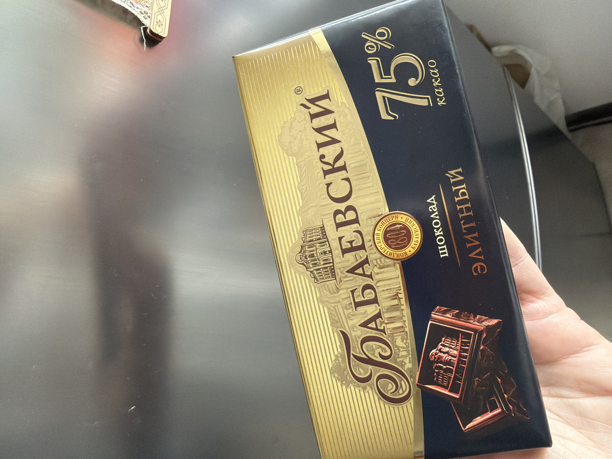 A hand holds a dark blue and gold chocolate bar in front of a metallic surface. The chocolate bar has the brand name "Бабаевский" in large, ornate golden lettering. Below this, it reads "ШОКОЛАД" and "ЭЛИТНЫЙ". To the right, the percentage "75%" is prominently displayed in gold, followed by "какао". There is a circular emblem with "1804" inside. The background appears to be a stainless steel appliance, possibly a refrigerator or a range hood, reflecting light. The scene is indoors, with no discernible external elements or location cues that would suggest the city of Khimki, Russia.