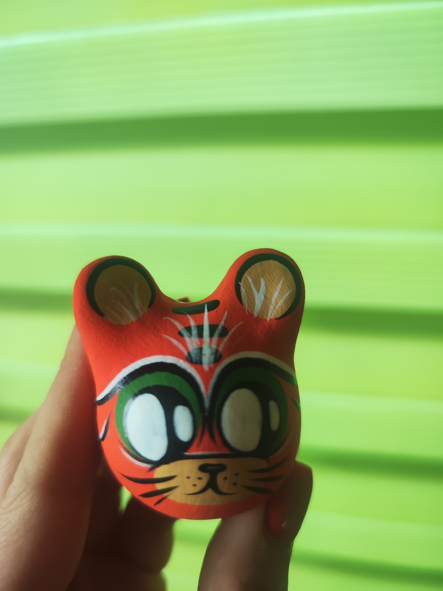 A close-up shot captures a small, hand-painted figurine of an animal head, likely a tiger or cat, held by a person's hand. The figurine is vibrant orange with distinct facial features painted in black, white, green, and a lighter orange/yellow for the snout and inner ears. It features large, round white eyes with black pupils, outlined by bright green circles. Black stripes are painted on its forehead and around its ears. A small black nose and mouth are visible on the lighter orange snout, with black lines extending outwards like whiskers.
A person's fingers gently cradle the figurine from below, with a hint of red nail polish visible on one fingernail. The background is a brightly lit, blurred, vibrant green surface, showing subtle horizontal lines that suggest a ribbed texture, possibly blinds or a panel, and creating a soft, out-of-focus effect. The lighting is bright and even, highlighting the colors of the figurine. No text is visible in the image.