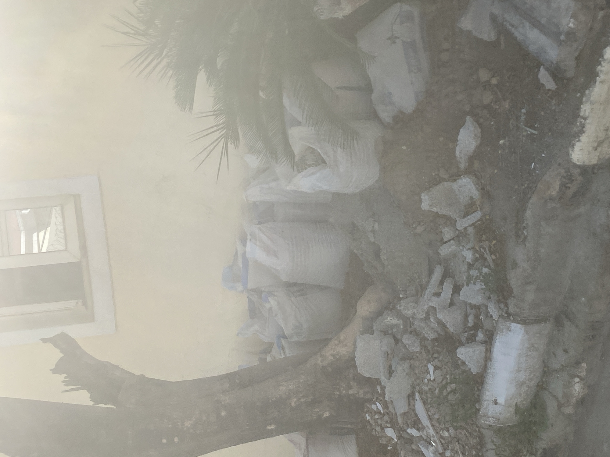 The image shows a scene in Gosa, Nigeria. A partially visible building with a window is on the left. Next to the building, there are several white sacks piled on top of each other. A bare tree branch lies across the bottom of the image. Debris and scattered pieces of concrete are visible on the ground near the sacks. The image has a hazy or dusty quality.