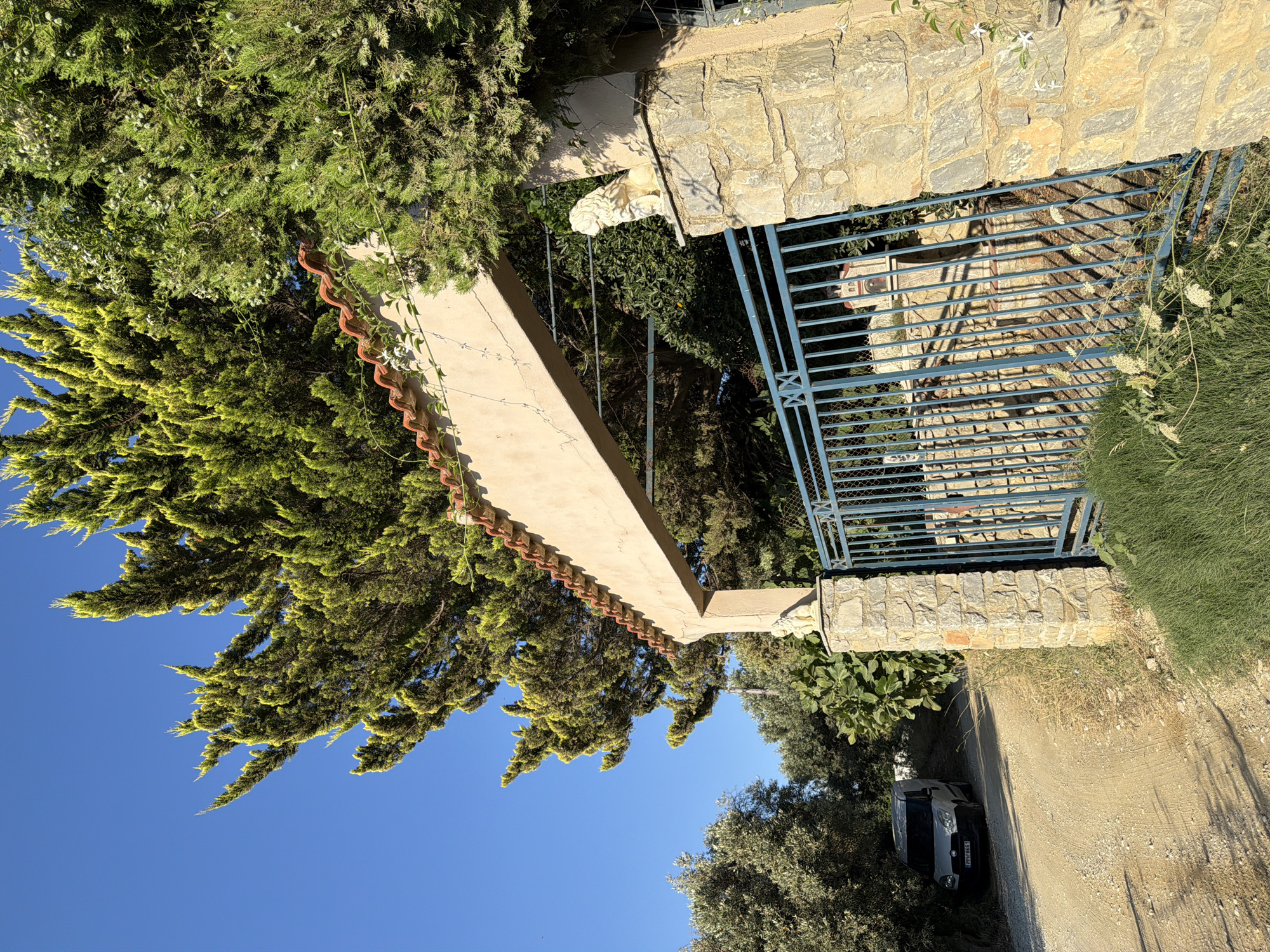 The image depicts an entryway to a property on the island of Skiathos, Greece. The entryway features a stone pillar topped with a white lion statue, supporting a stucco archway adorned with terracotta roof tiles. A blue metal gate stands open, revealing a glimpse of a small building within the property. To the left of the entryway, a dirt road leads past a white van parked beside a cluster of trees. The overall scene is bathed in the warm, clear light of a sunny day.