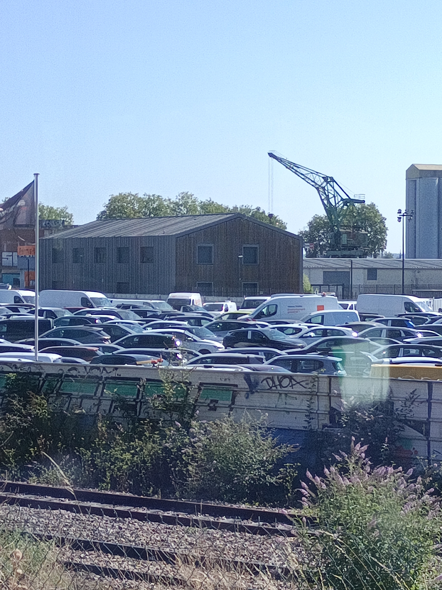 The image depicts a parking lot filled with numerous cars. A large industrial crane is visible in the background, suggesting a port or industrial area. Several buildings, including a warehouse-like structure, are also present. The sky is clear and blue. The foreground shows railway tracks and vegetation. The scene is likely located in the port area of Rouen, France, given the presence of industrial elements like the crane and buildings often found in such locations. There are no people visible in the image, and the cars are stationary.