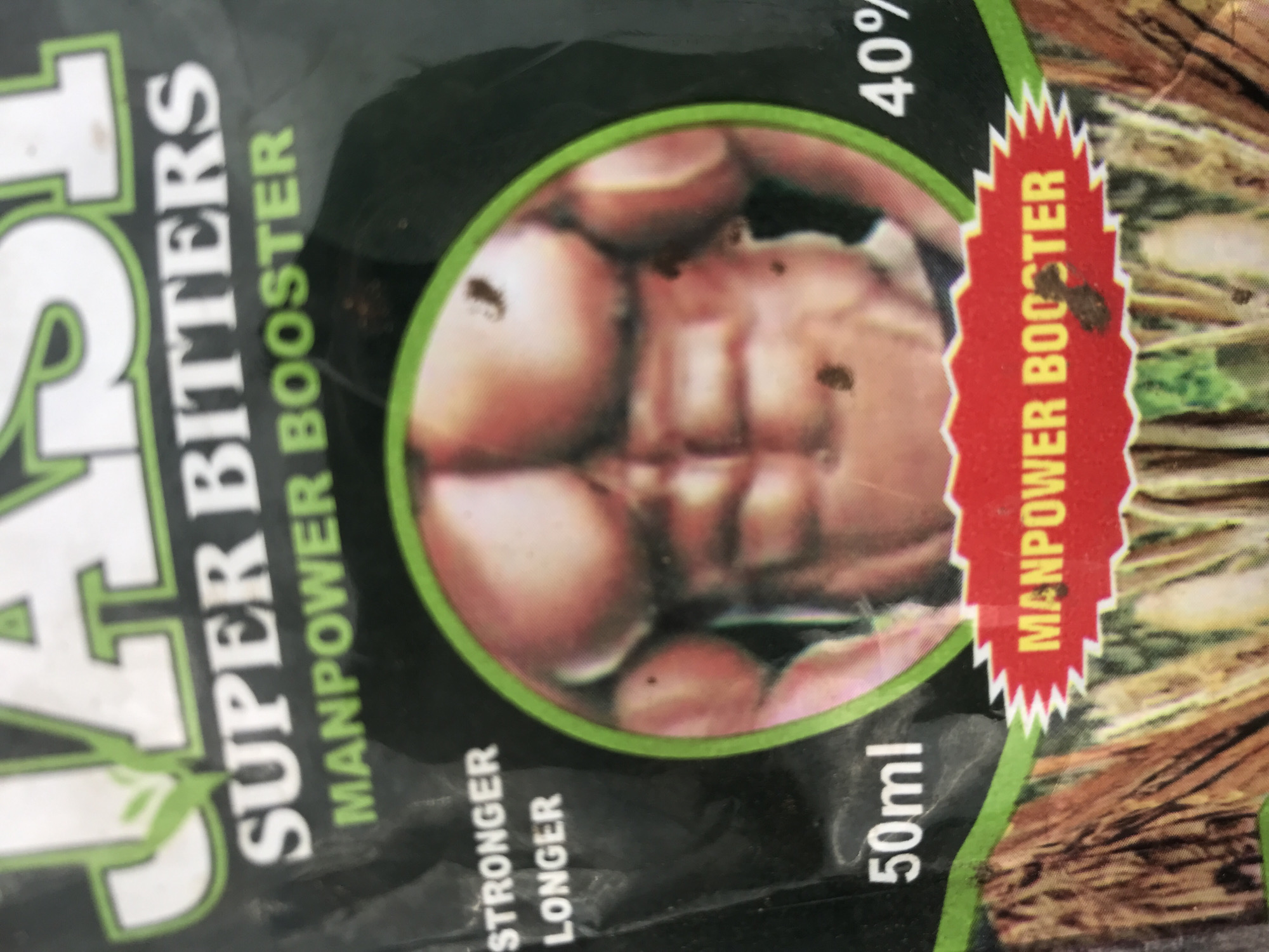 A product label, likely for a dietary supplement or beverage, is depicted in close-up. The top portion of the label displays the brand name, partially visible as "JASH" in large white letters with a green outline. Below this, the phrase "SUPER BITTERS" is printed in white capital letters with a green outline, followed by "MANPOWER BOOSTER" in green capital letters.
Central to the label is a circular graphic featuring a light-skinned, highly muscular male torso, showcasing well-defined pectoral muscles and a prominent six-pack abdomen. Several small, dark, irregular specks, consistent with debris or dirt, are present on the surface of this graphic. To the left of the circular graphic, the words "STRONGER" and "LONGER" are stacked vertically in white capital letters. Below the graphic, "50ml" is printed in white. To the right, "40%" is visible in white. The bottom of the label features a jagged-edged red banner with "MANPOWER BOOSTER" in yellow capital letters. The lower background of the label shows an image of intertwining, gnarled tree roots or wood. The packaging material appears to be flexible, with minor creasing. This product packaging is situated in the city of Rafin Par, Nigeria.