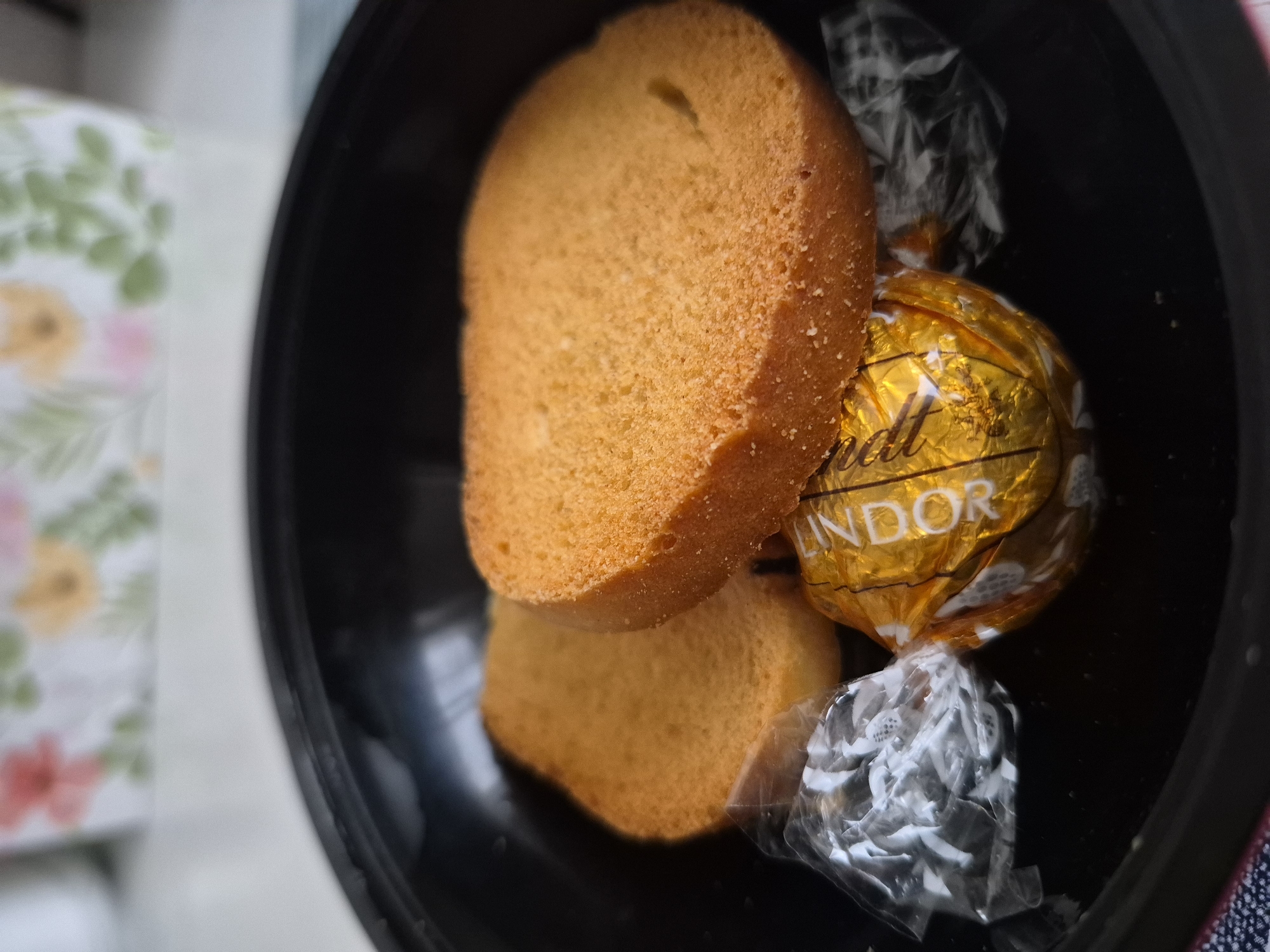 In an overhead shot of a black bowl, two slices of golden-brown toast are arranged next to a foil-wrapped piece of chocolate. The toast appears to be slightly stale, with a rough, grainy texture on its surface. One slice is positioned slightly in front of the other, with the front slice tilted upwards, revealing its textured underside. Nestled between the toast slices and to the front is a gold foil-wrapped chocolate, its wrapper crinkled and imprinted with the "LINDOR" logo in dark lettering. To the left of the chocolate is a clear plastic wrapper, also crinkled, which likely contained another item that has been removed or is obscured. The background of the image is blurred, showing a white surface with a floral pattern. The scene appears to be indoors, possibly on a table.
