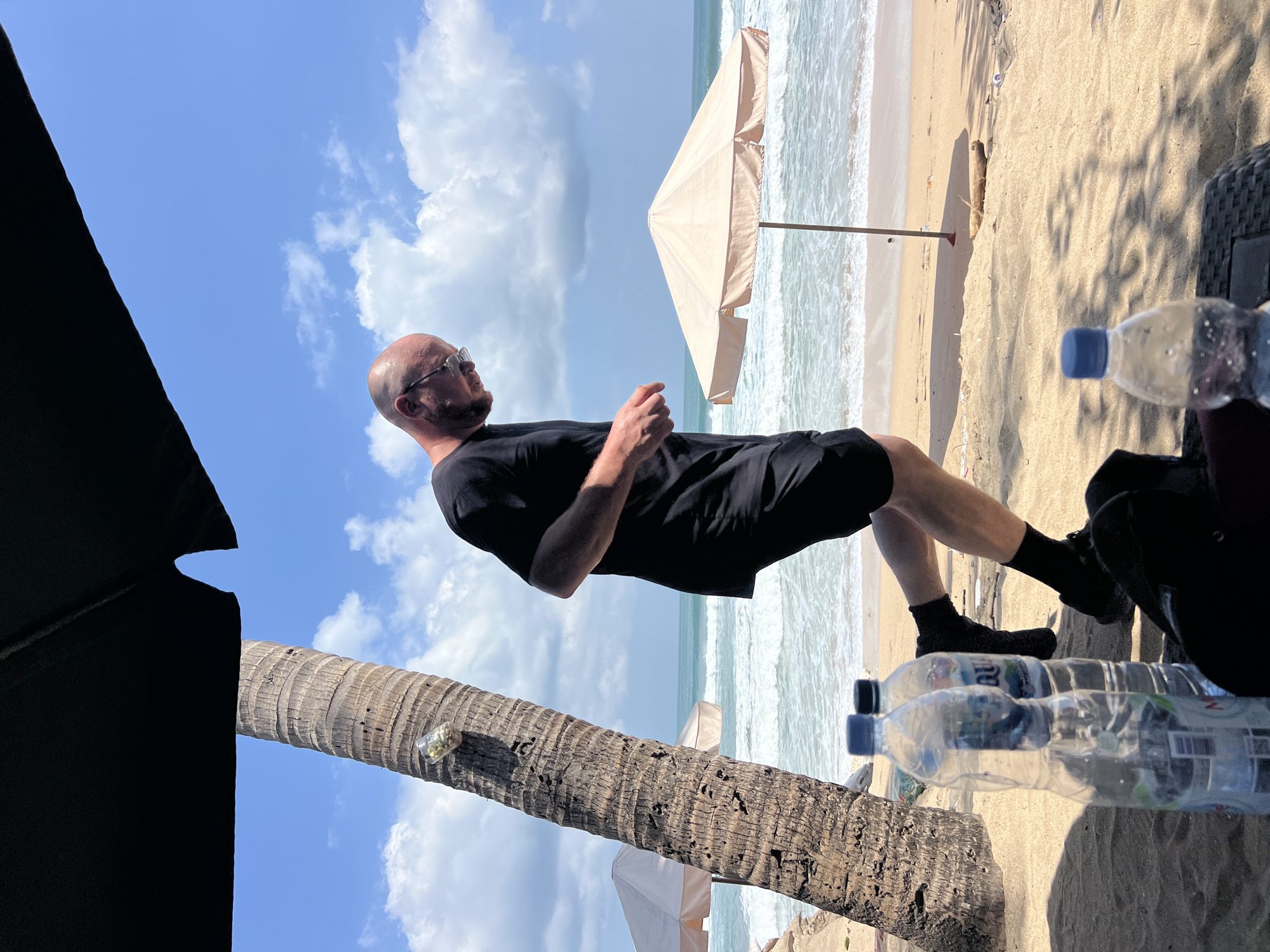 A bald man wearing sunglasses, a black t-shirt, and black shorts runs along a sandy beach in Kuta, Indonesia. The man is mid-stride, with his left leg forward and his right arm bent at the elbow. He is looking down and to his left. Behind him, the ocean waves crash onto the shore under a blue sky with white clouds. Two beach umbrellas, one white and one beige, are visible in the background. A palm tree stands to the left of the man, and a few plastic water bottles are on the sand in the foreground. The scene suggests a recreational activity on a sunny day.
