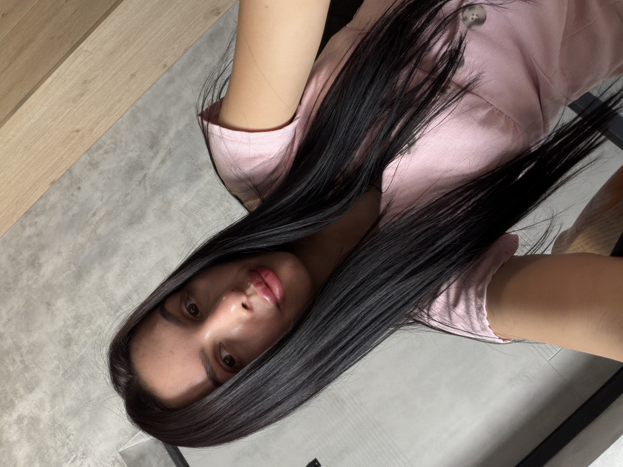 A young woman with long, dark hair and light skin is pictured in a selfie. She is wearing a pink collared shirt. The background appears to be an indoor setting with a textured grey wall and a wooden surface.