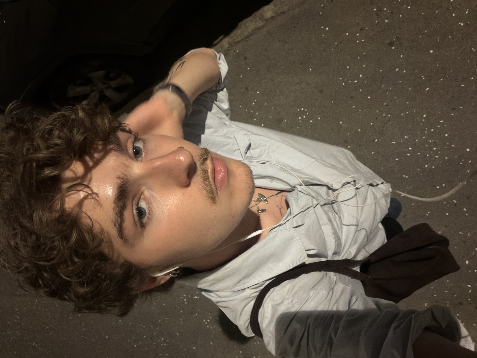 A young person with curly brown hair and light blue eyes, wearing a light blue button-down shirt and white earbuds, stands on a sidewalk at night. The person has a thin mustache and a nose ring. A dark brown bag is slung over their shoulder. In the background, the wheel and dark silhouette of a car are visible. The sidewalk is a dark gray asphalt surface with scattered white debris. The scene is illuminated by artificial light, casting shadows. The location is Vračar Urban Municipality, Serbia.