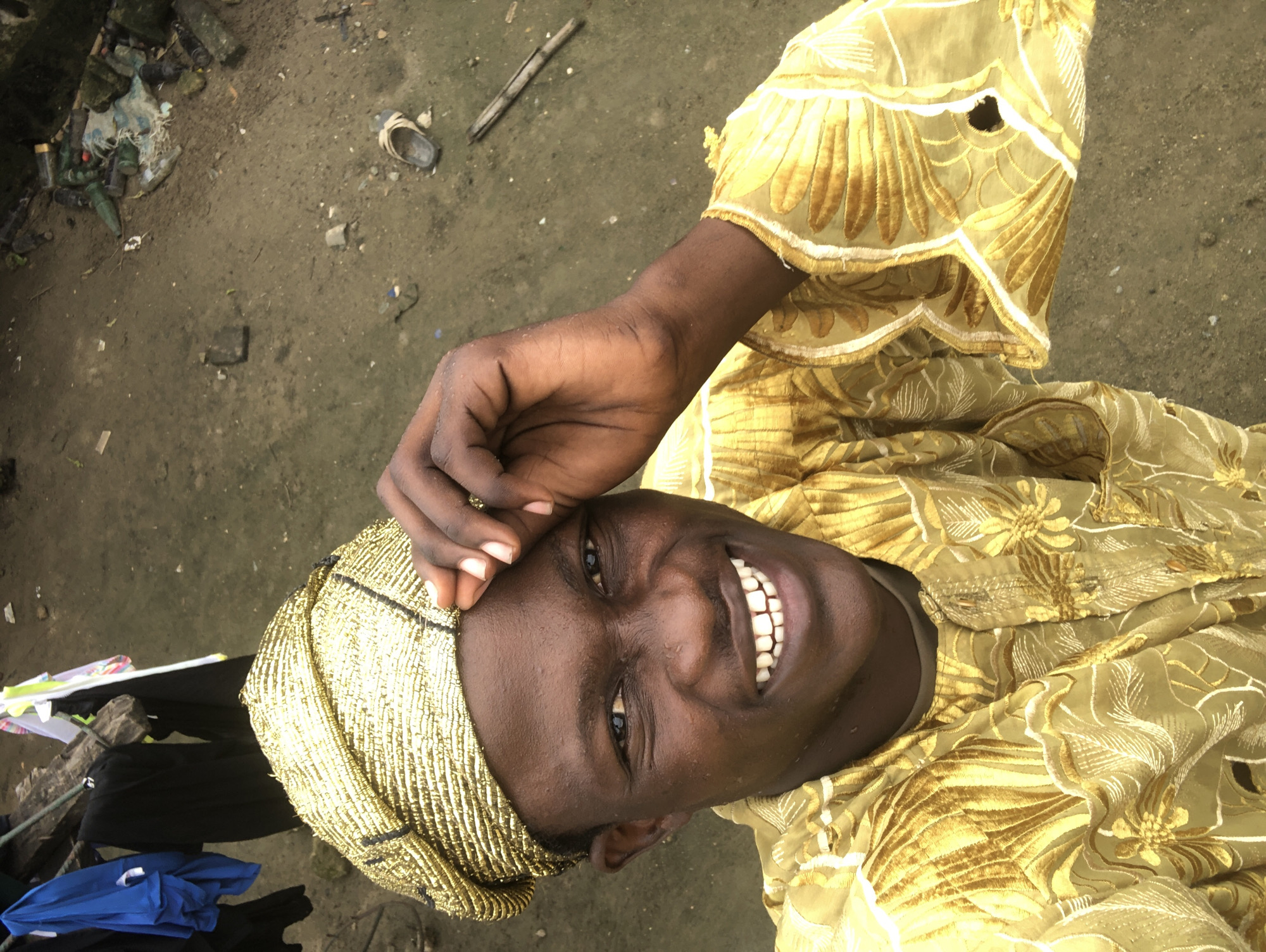 A young Black male is posing for a selfie outdoors. He is wearing a golden, patterned outfit and a matching headwrap. His right hand is touching his forehead. The ground beneath him appears to be dirt or mud, with scattered debris including plastic bottles, rocks, and a worn sandal. In the background, there is a wooden structure with clothes hanging from it, including a blue shirt and black garments. This scene is likely in Ado, Nigeria, given the environmental context which suggests a less developed area.