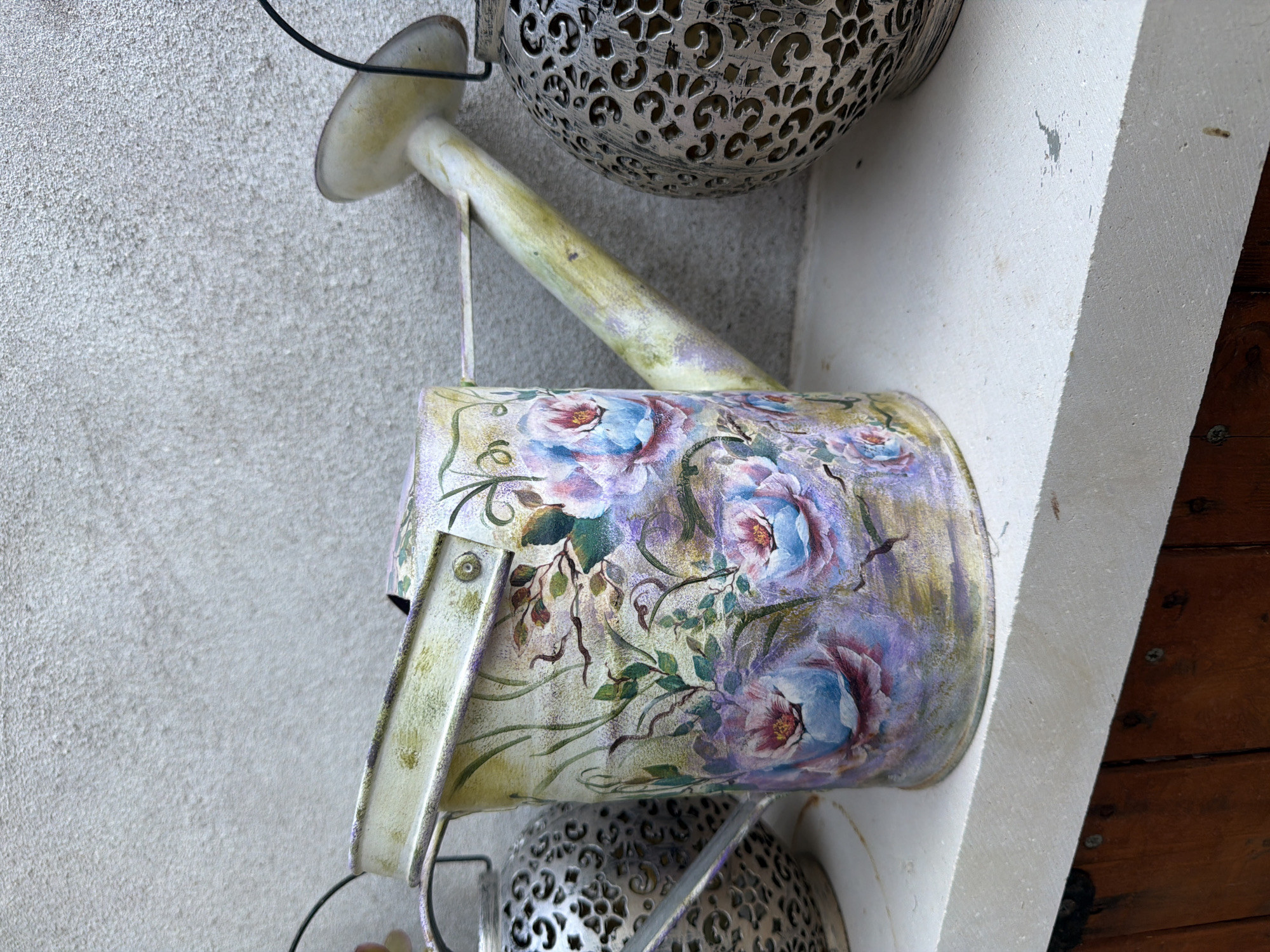 This image features a decorative watering can adorned with a floral motif. The watering can, painted with roses in shades of pink, purple, and blue, hangs from a wire. It is positioned against a textured, light-colored wall. In the background, a portion of an ornate, silver-colored lantern with intricate cutouts is visible, also hanging. The watering can itself appears to be made of metal and shows signs of weathering or a deliberately distressed finish, with areas of green and gold patina. The spout of the watering can extends to the left. To the right of the watering can, a light-colored, angular structure, possibly part of a building or furniture, juts out. The overall setting suggests an outdoor or semi-outdoor decorative display, possibly in a garden or patio area. The lighting suggests it is daytime, with no strong shadows indicating a specific time. The style of the decor and the presence of the lantern and watering can evoke a charming, perhaps Mediterranean, aesthetic. No people are visible in the image. The setting appears to be a wall in Laganas Municipal Unit, Greece, judging by the provided location context.