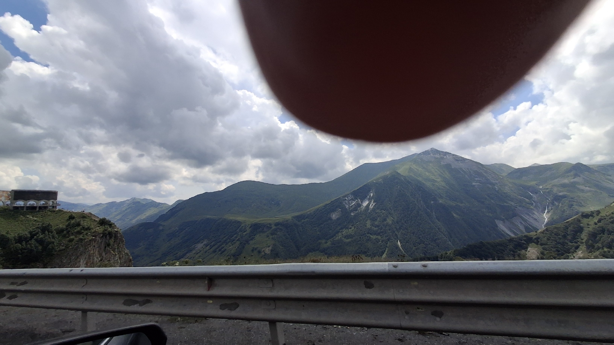 The image shows a mountain landscape with a large, circular object obscuring part of the sky in the upper portion of the frame. In the middle ground, a building with arched openings sits on a rocky outcrop. Further in the background, rolling green mountains dominate the scene, with a waterfall visible cascading down one of the slopes. A metal guardrail runs across the lower part of the image, suggesting the vantage point is from a road. The sky is filled with dramatic, grey and white clouds. The scene appears to be a scenic viewpoint, possibly along a road in Gudauri, Georgia, known for its mountainous terrain and ski resorts.