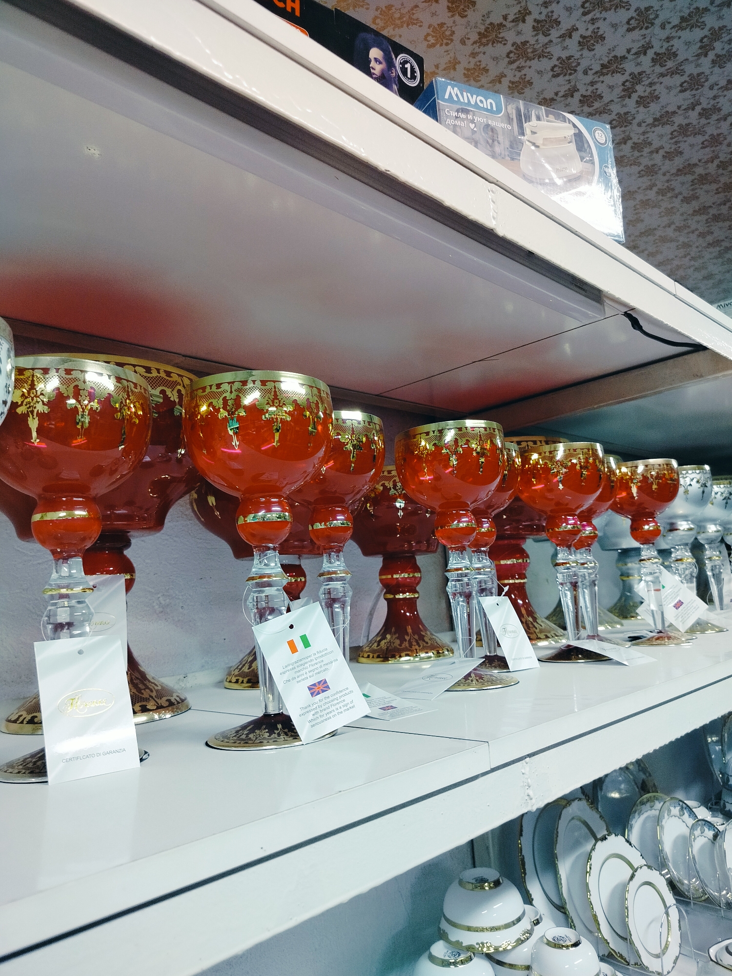 The image displays a retail setting, likely a shop, showcasing various glassware and dinnerware items. The focus is on a shelf filled with ornate, red glass goblets or vases with gold embellishments. These items have clear glass stems and bases, also accented with gold. Attached to some of the items are white tags with text and flags, including an Italian flag and a Union Jack, suggesting an international origin or marketing.
Below this shelf, another shelf is partially visible, displaying white porcelain plates with gold trim and stacked white porcelain cups and saucers, also adorned with gold detailing.
In the background, on a higher shelf, a box with "Mivan" branding and an image of a kettle is visible. The shop's walls appear to be decorated with a patterned wallpaper. The lighting suggests an indoor environment, possibly a store with display shelves. The overall impression is that of a well-stocked establishment selling decorative and functional household items. The location is specified as Көлкент, Kazakhstan.