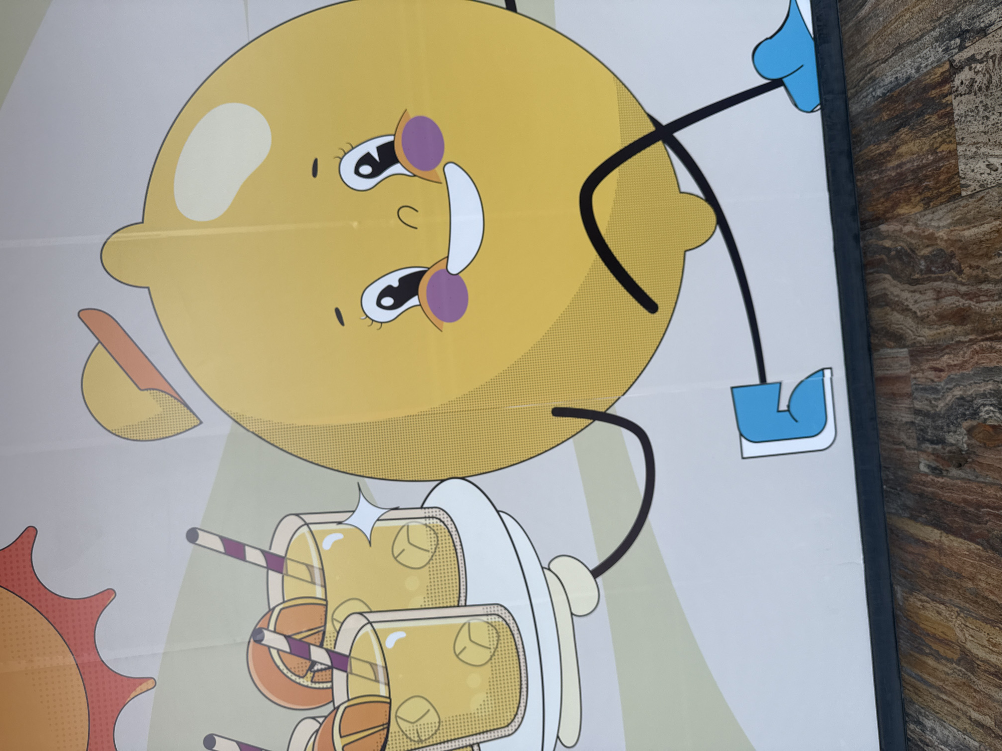 The image shows a close-up of a cartoon-style poster. The poster features a large yellow lemon character with a smiling face, complete with stylized eyes, cheeks, and a mouth. The lemon has arms and legs depicted as simple black lines with blue shoes. Below the lemon, there are cartoon glasses filled with yellow liquid and ice cubes, arranged on a white platter. To the left, a portion of a cartoon sun is visible. The background is a mix of light green and white. The poster appears to be framed in a dark border, and a textured surface is visible on the right side of the frame. This poster is located in Mexico City, Mexico.