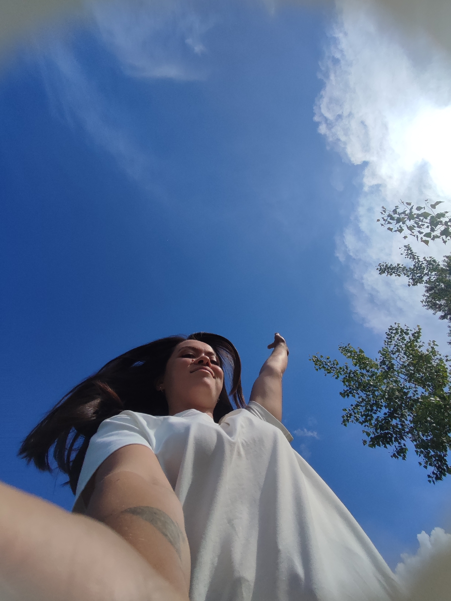 A low-angle shot shows a woman in a white t-shirt with her arm extended toward a tree. The sky is blue with scattered clouds. A tattoo is visible on her forearm. The location is plausibly in the city of Chaikovsky, Russia, based on the image itself.