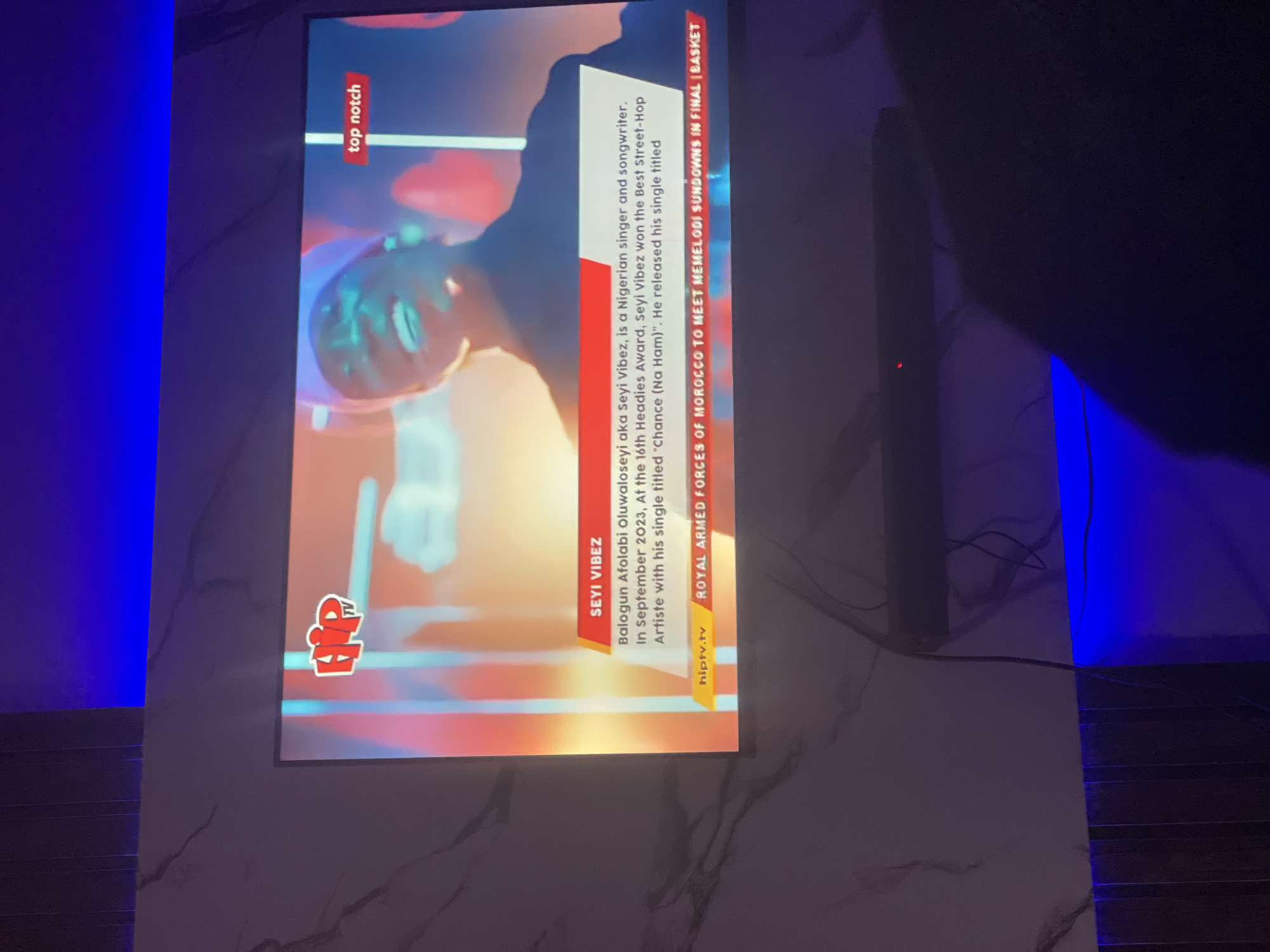 The image shows a television screen mounted on a wall with a marble-like pattern. The TV displays information about Seyi Vibez, a Nigerian singer and songwriter, with text describing his achievements. A "top notch" banner is visible in the upper left corner of the screen. At the bottom, there's a banner about the Royal Armed Forces of Morocco meeting Mamelodi Sundowns in the final of a basket. The bottom left of the screen displays the hiptv.tv logo. To the right of the screen, an unidentifiable object and its associated cables are visible. The wall surface has a marble-like pattern, and there is a light source emitting blue light. It is unclear if this is in Lokoja, Nigeria.