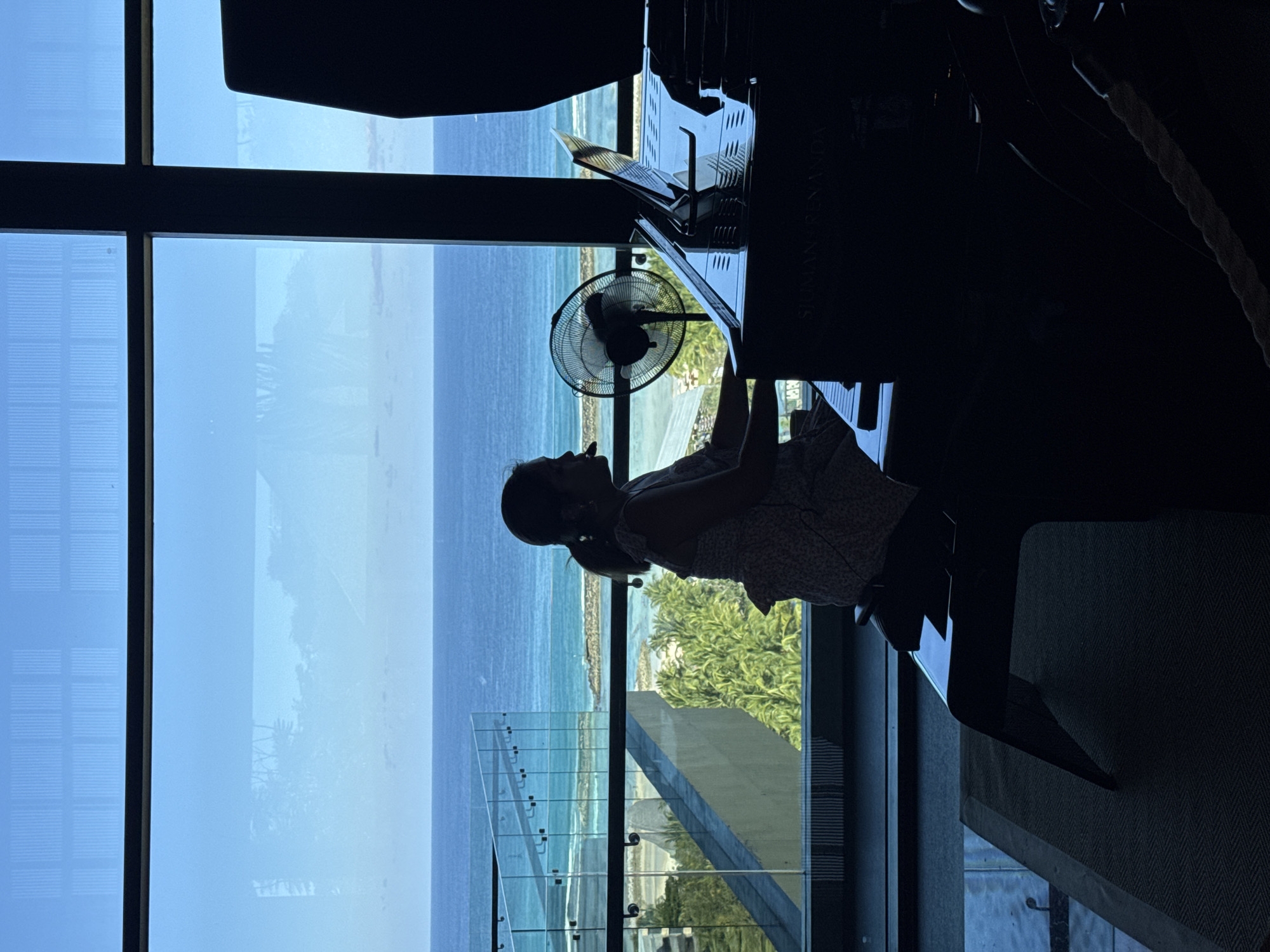 A person is silhouetted while playing a piano indoors near a large window overlooking the ocean in Kuta Selatan, Indonesia. The ocean view includes a beach, palm trees, and a partially visible structure. A fan sits on top of the piano, behind the person. The view from the window suggests a high-rise building.