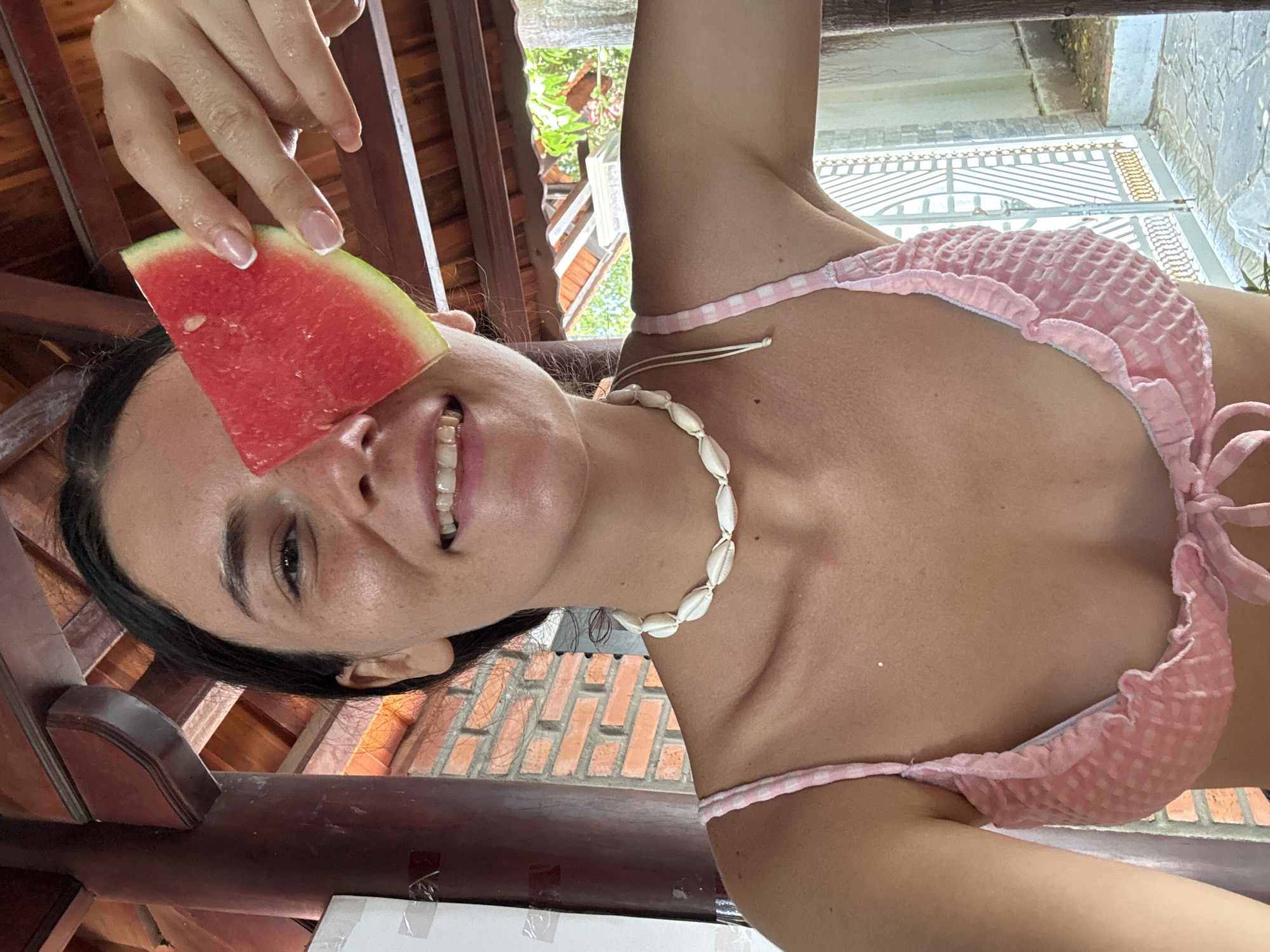 A young woman, positioned slightly off-center to the right, is seen from the chest up, holding a slice of watermelon over her right eye. She is smiling with her mouth open, revealing her teeth. She wears a pink, checkered bikini top with ruffled edges and a tie in the center. Around her neck, she has a white shell necklace. Her left arm is visible, and her hand is resting on her upper chest.
The background shows a wooden ceiling with visible beams, a red brick wall, and a glimpse of greenery and a decorative metal gate. The setting appears to be an outdoor or semi-outdoor area, possibly a cafe or restaurant with a traditional architectural style common in Mũi Né, Vietnam. The lighting suggests it is daytime.