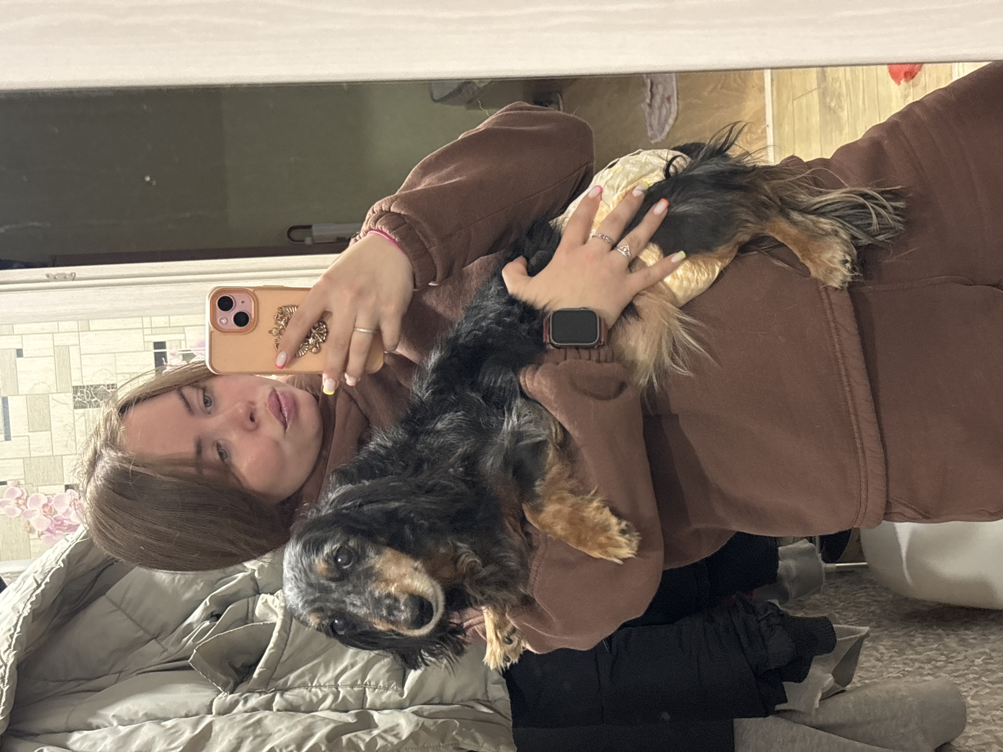 A young woman is holding a black and tan long-haired dachshund and taking a selfie. She is wearing a brown sweatsuit. She is wearing a watch on her left wrist and several rings on both hands. The woman is holding a cell phone in her right hand. The background is a bedroom with bedding, curtains, and other objects. There is no evidence to suggest that this photo was taken in Izhevsk, Russia.