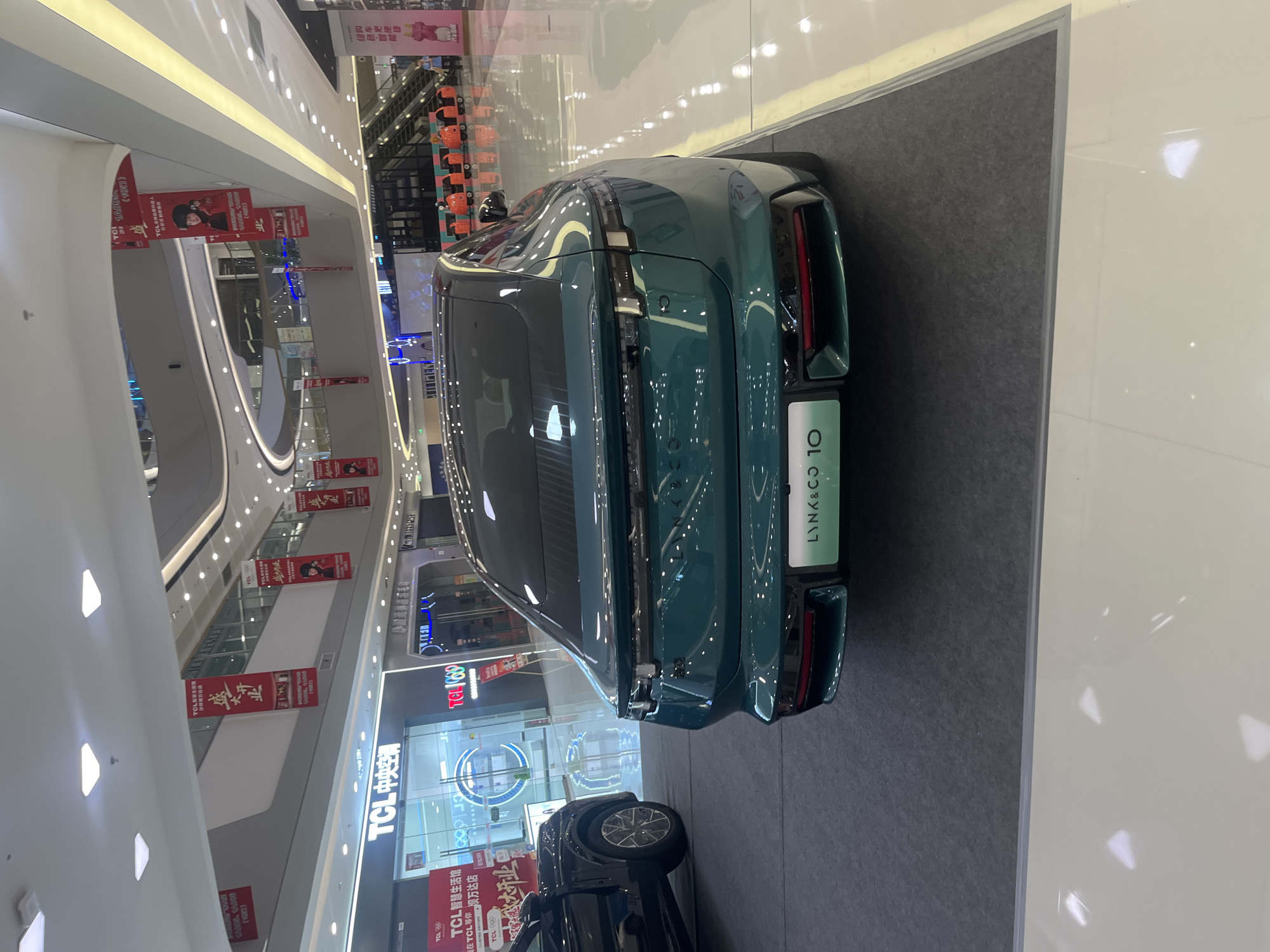 A dark green Lynk & Co 10 sedan is parked on a gray carpet in a brightly lit indoor shopping mall. The car's rear is facing the camera, showcasing its taillights and the "LYNK&CO 10" badging. To the left of the sedan, part of a black SUV is visible. In the background, a TCL electronics store and other shop fronts are apparent. Banners with Chinese text are hanging from the ceiling. The mall's architecture features multiple levels, escalators, and a polished floor reflecting the overhead lights. The scene appears to be within the Chongqing Global Shopping Center in Shapingba District, China, given the presence of TCL stores and the general mall aesthetic.