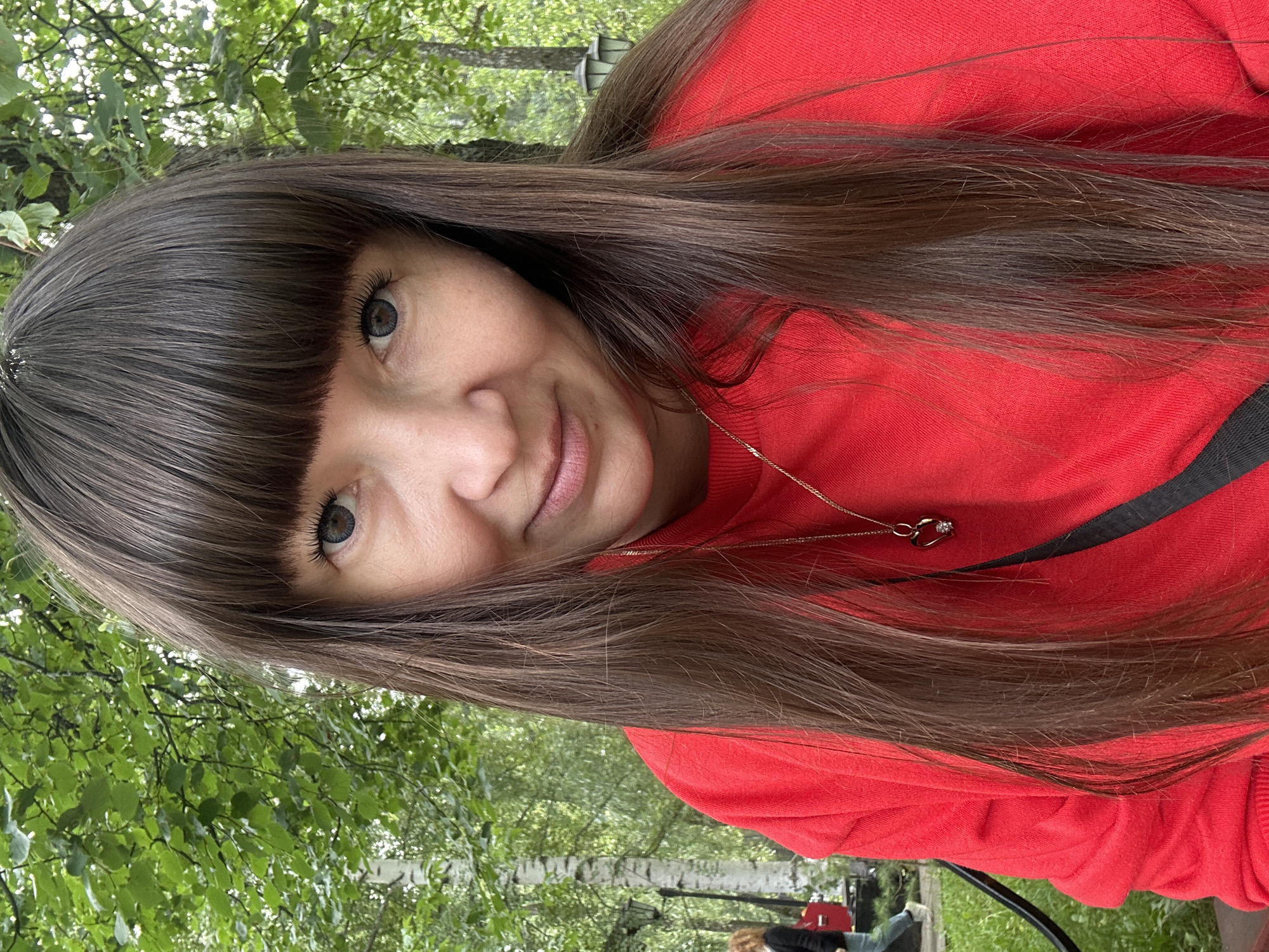 A medium shot photograph shows a woman with long, dark brown hair and bangs, wearing a red sweatshirt and a gold necklace with a heart-shaped pendant. She is outdoors in what appears to be a park, with lush green trees and foliage in the background. A person can be seen blurred in the distance to the left of the frame, and lampposts are visible among the trees. The lighting suggests it is daytime, possibly overcast, contributing to the soft, natural look of the scene. The woman's facial expression is gentle and she is looking directly at the camera. The setting suggests a relaxed, casual atmosphere, possibly within the city of Chaikovsky, Russia, given the presence of birch trees which are common in Russian landscapes.