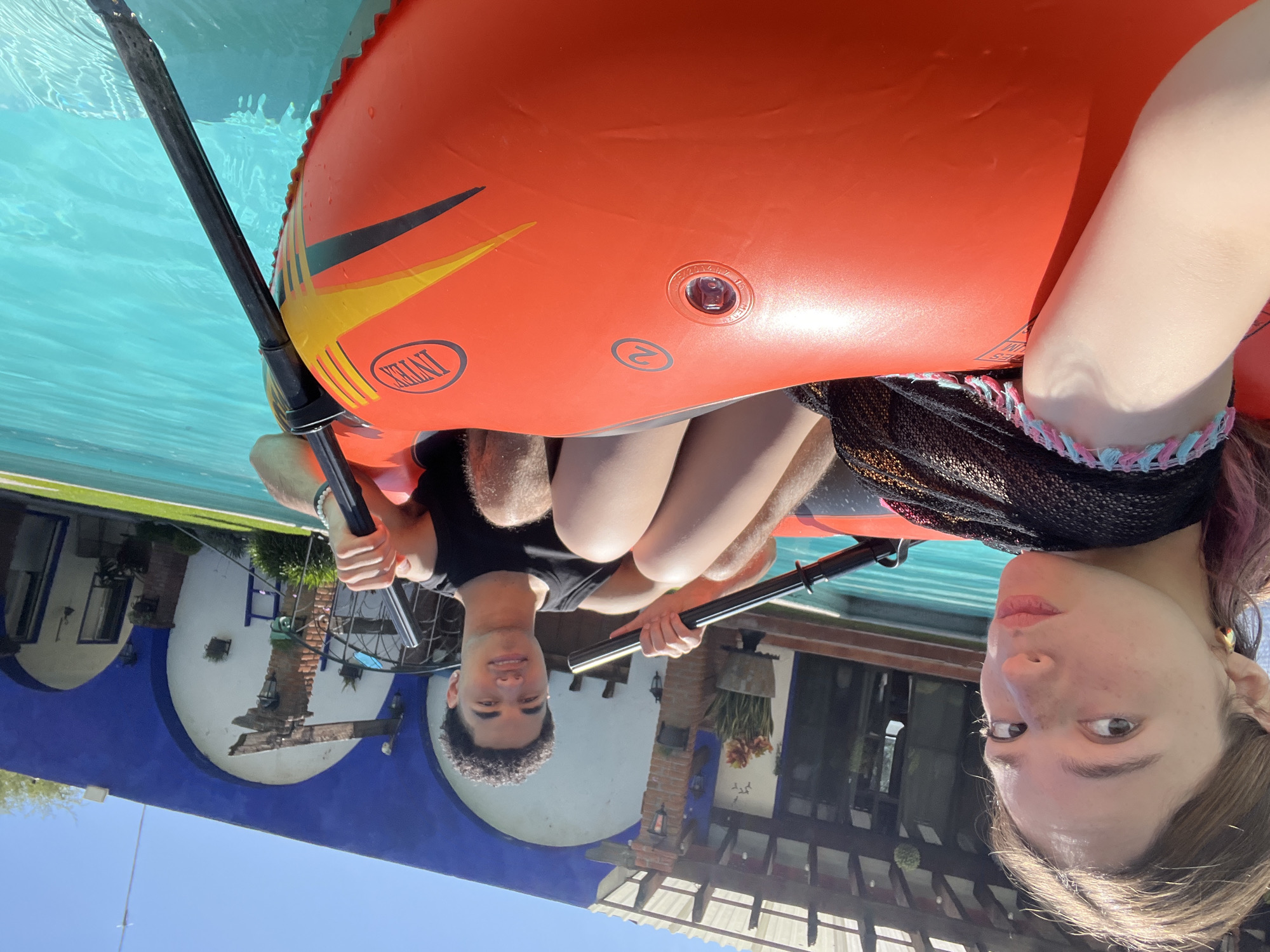 Two people are in an orange inflatable raft in a swimming pool. One is a young man with curly hair, wearing a black t-shirt. The other is a young woman with light hair, wearing a black top with a colorful wristband. They are each holding an oar. The pool is surrounded by buildings with a blue exterior wall. The location is Puebla, Mexico.