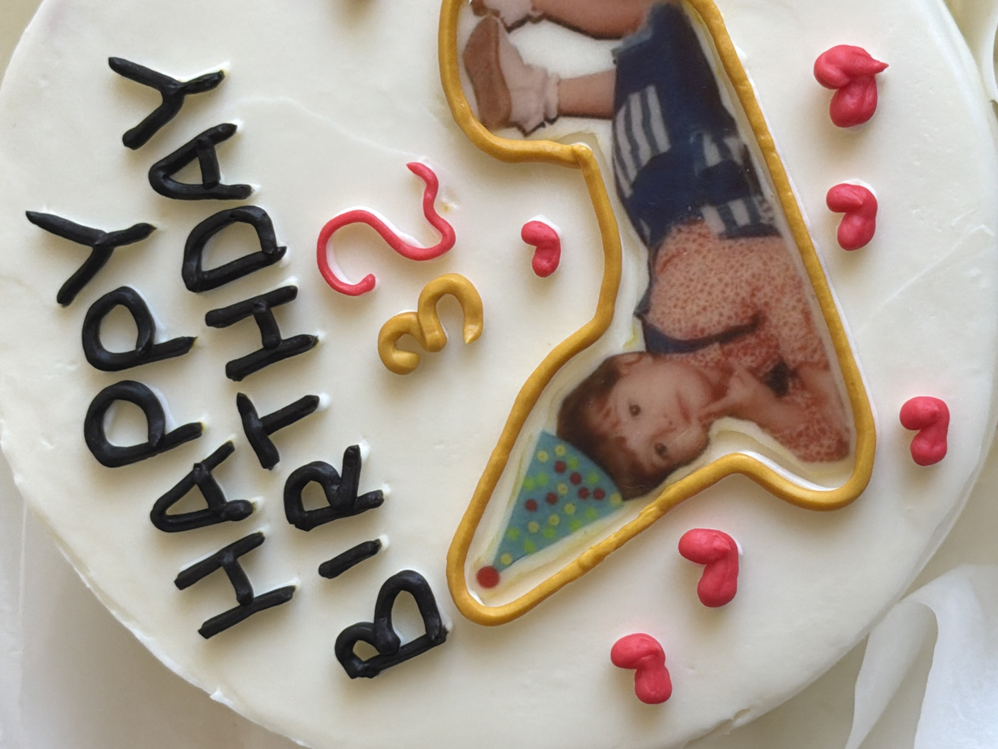 The image shows a close-up of a decorated cake, presumably for a birthday. The cake has white frosting and is decorated with black lettering that reads "HAPPY BIRTHDAY". To the right of the lettering, there are red and gold numbers, possibly representing an age (23). There's also a framed edible image of a child wearing a party hat, and small red candy decorations scattered around the image. The cake's location is not discernible from the image.