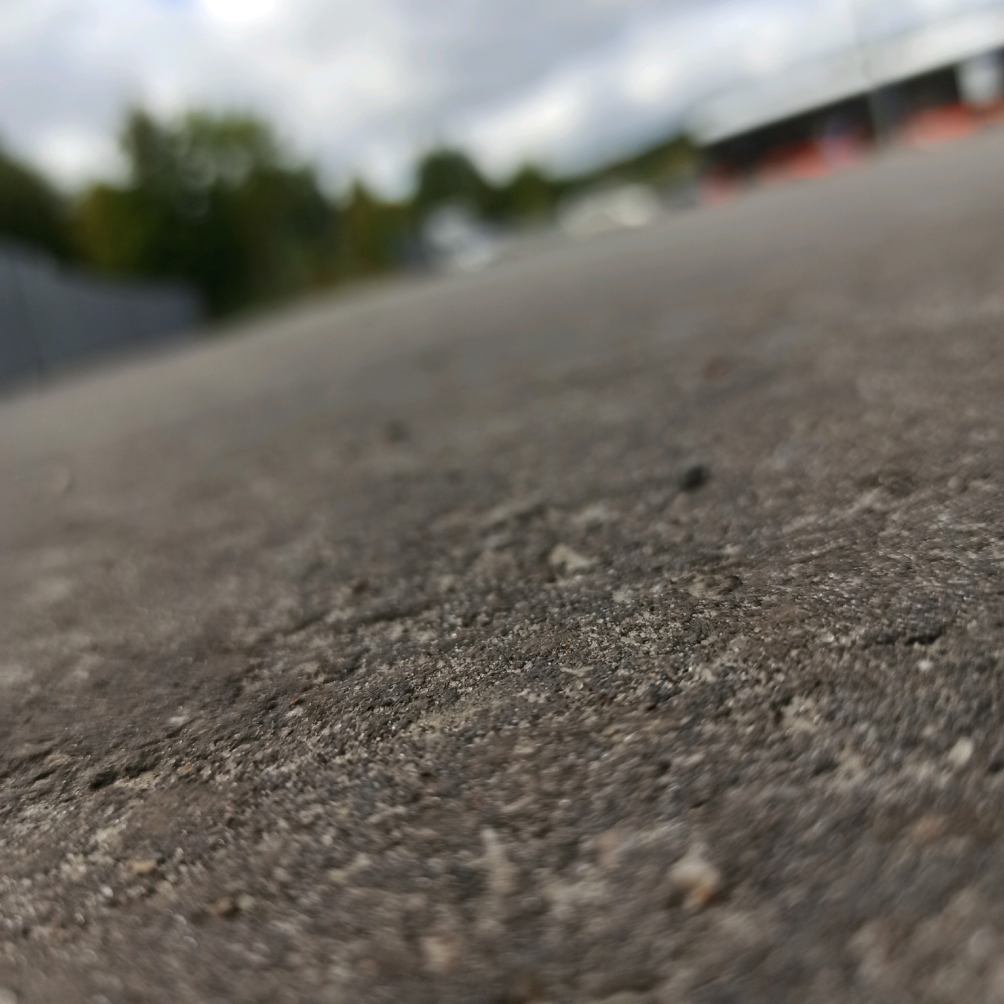 The image is a close-up, low-angle shot of a textured, gray surface, likely asphalt or concrete, with small pebbles and debris. The foreground is in sharp focus, revealing the granular texture of the ground. The background is out of focus, showing blurred shapes of green trees and a structure with an orange element, possibly a building or a sports facility, under a cloudy sky. The scene does not contain any people or discernible actions. There is no information in the image that would allow for the determination of its location in the city of Nizhny Novgorod, Russia.
