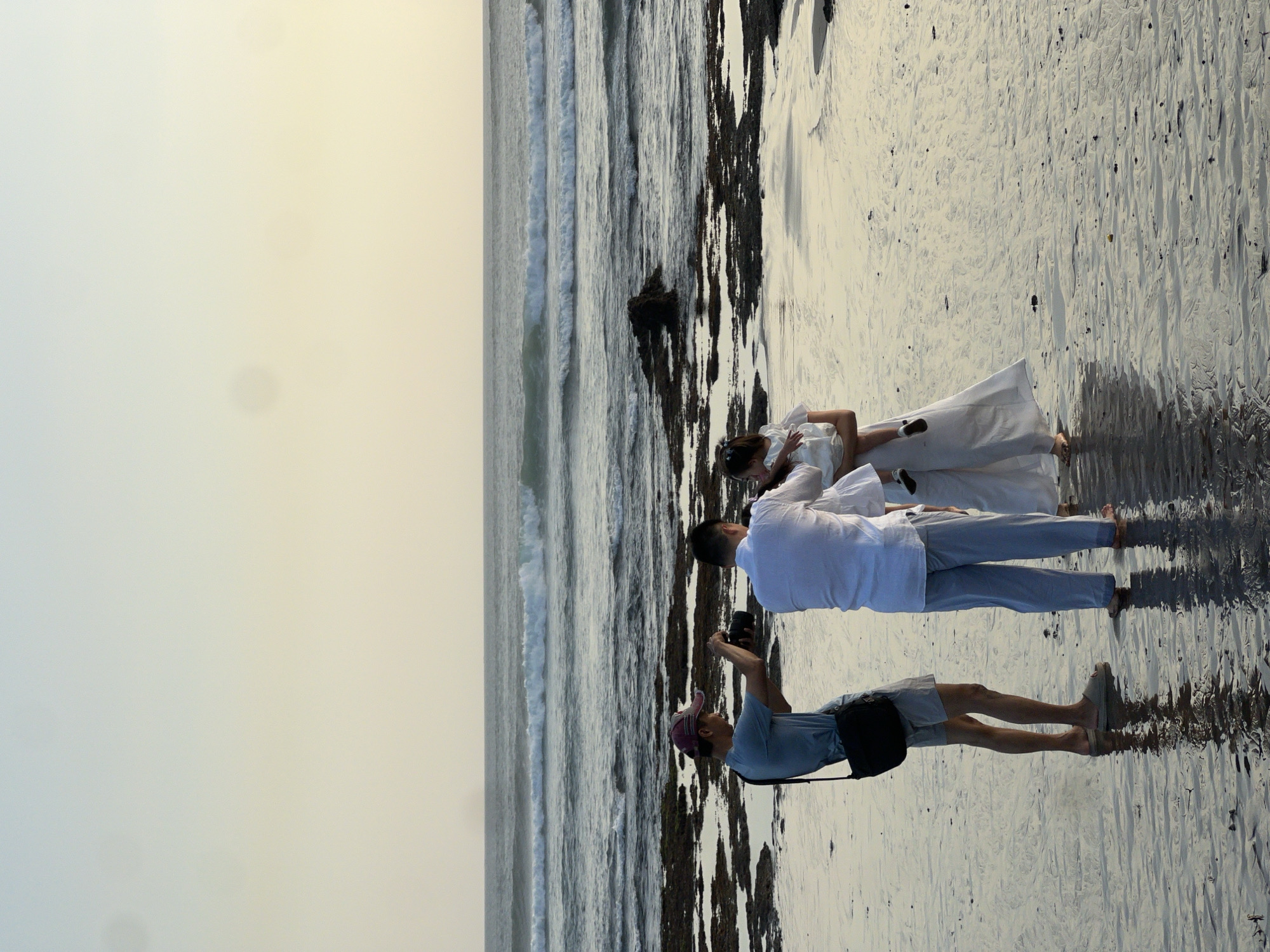 A photographer in a light blue t-shirt and light grey shorts with a black bag slung over his shoulder stands on a wet sandy beach, holding a camera and appearing to take a photo. To his right, a man in a white long-sleeved shirt and light blue pants is holding a young child, who is wearing a white dress. The man in white is looking down at the child, and the photographer is looking at them. They are positioned on a beach with rocks and shallow water. The ocean is in the background, with waves rolling in, and the sky is a pale yellow and blue, suggesting it is either sunrise or sunset. The scene appears to be in North Kuta, Indonesia.