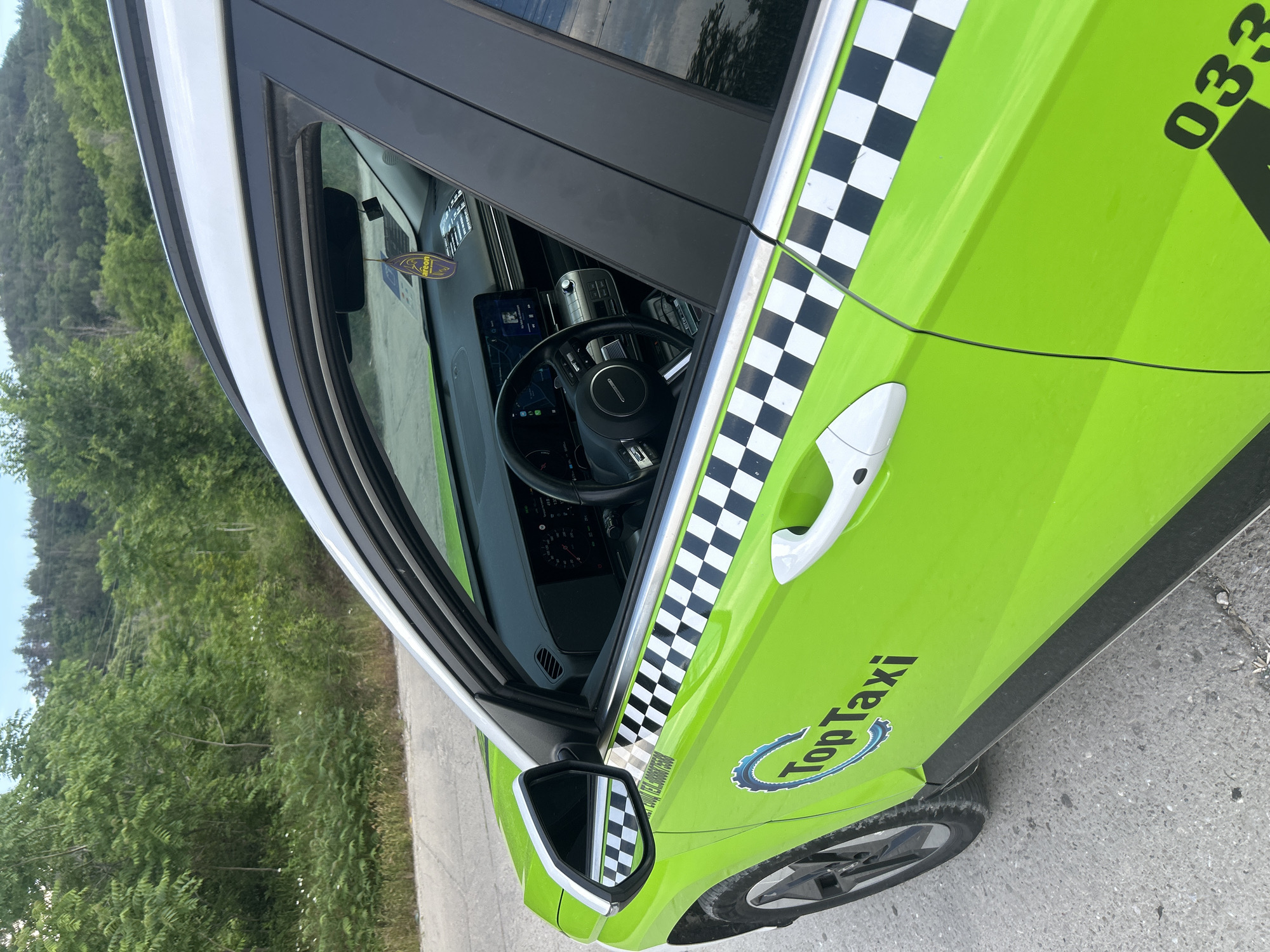 An outdoor, eye-level shot captures the side of a bright lime green and white taxi, positioned at an angle with the front of the car facing towards the viewer's right. The car features a white roof and a prominent black and white checkerboard strip running horizontally along the side, just below the windows. The main body of the car is a vibrant lime green. White door handles are visible on the passenger side.
Through the open window, part of the car's interior is visible, including a dark-colored steering wheel with a central logo, a digital dashboard display, and other controls. An "AREON" brand air freshener hangs from the rearview mirror inside the cabin. The exterior rearview mirror, white on its outer casing, reflects parts of the car's side and the surrounding environment.
The taxi is parked or stationary on a paved surface, likely concrete or asphalt, with dense green foliage, including trees and bushes, forming the background. The natural light suggests it is daytime, under clear weather conditions.
Visible text on the side of the green car includes "Top Taxi" with a blue gear-like logo underneath, and a large black number "033" positioned towards the rear of the car. Some smaller, illegible text is reflected in the exterior mirror, possibly a phone number starting with "TEL:".