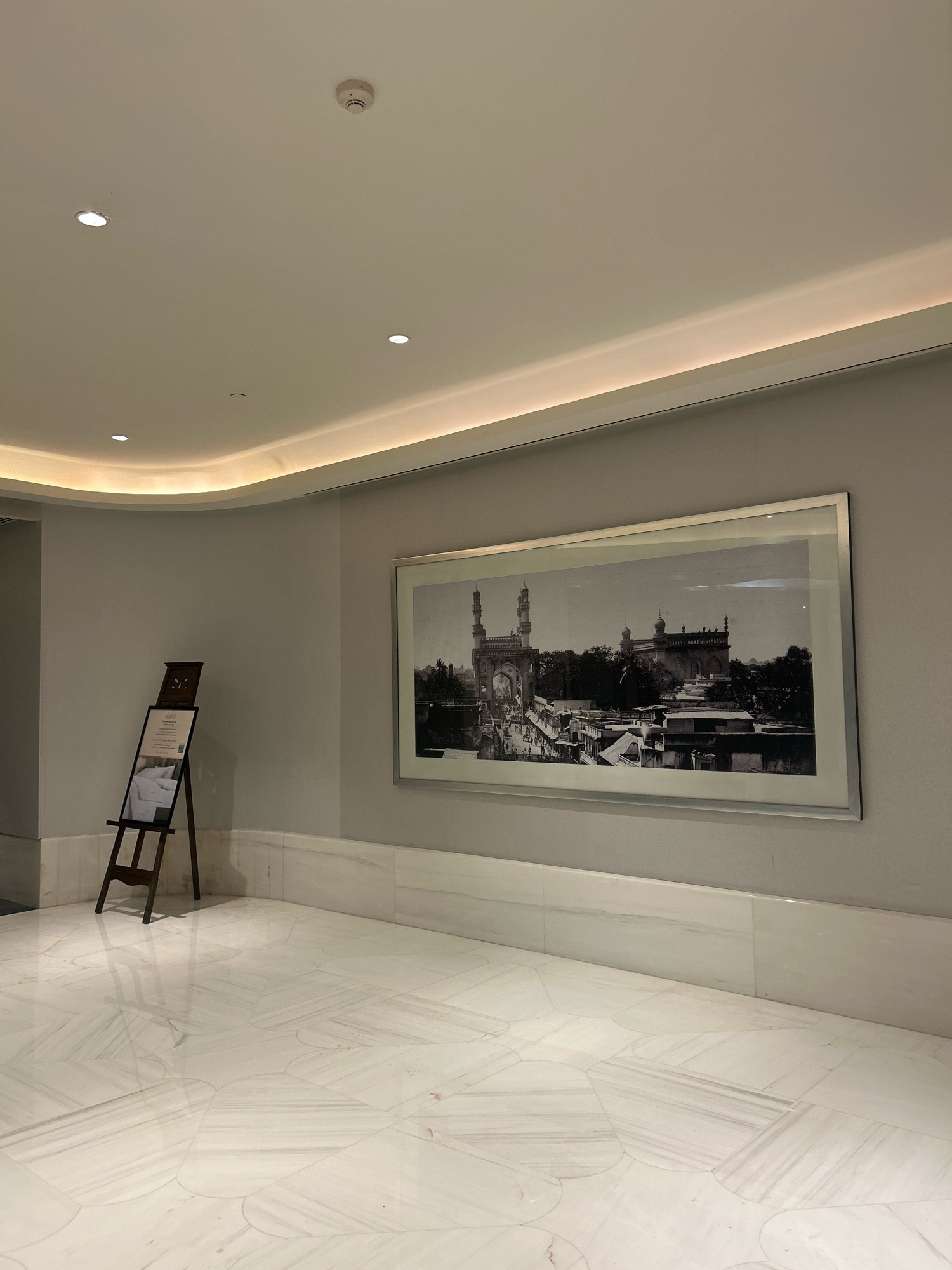 The image shows a wide, well-lit interior hallway with marble flooring. On the left, an easel displays a framed document with images of bedding. To the right, a large framed black and white photograph depicts the Charminar in Hyderabad, India. The photograph captures the iconic monument with surrounding buildings and bustling activity. The hallway features a modern, minimalist design with recessed lighting in the ceiling and a cove light running along the wall. The overall impression is one of a clean, elegant, and contemporary space.