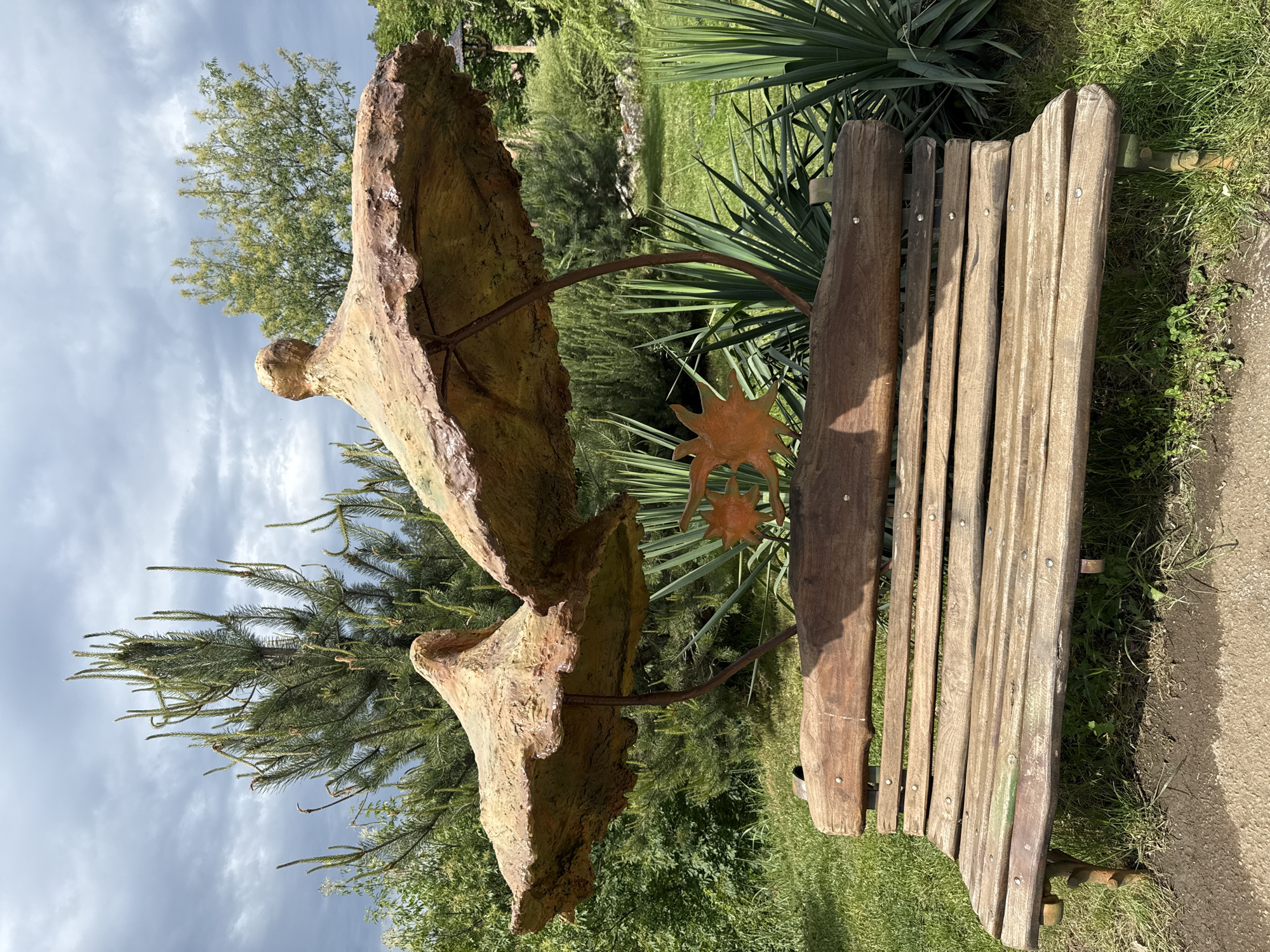 The image shows a wooden bench with a sculptural addition in a grassy area, likely a park or garden. The bench is made of weathered planks, with a dark wooden support structure. A stylized sculpture is integrated with the bench, featuring two tan, irregular, shell-like forms connected by a metal rod. A metal sun-shaped ornament is attached between the sculpture and the bench. The background includes various greenery, including trees and shrubs. Based on the prompt, the location is likely Lukovit, Bulgaria. No people are visible.
