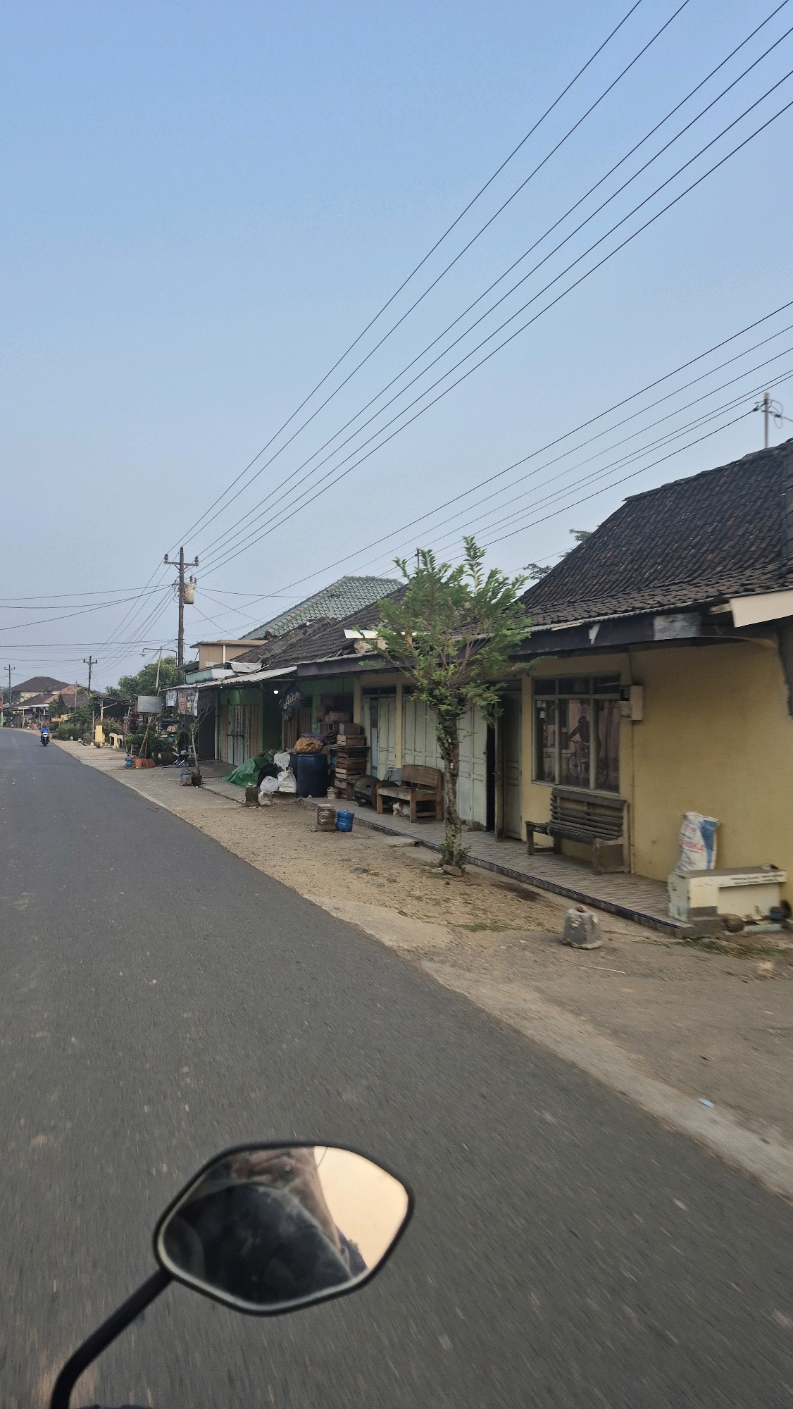 The image captures a view from a motorcycle traveling along a paved road in Wiroko, Indonesia, under a clear, pale blue sky. Overhead, multiple power lines stretch across the frame, supported by utility poles. The road runs parallel to a row of small, single-story buildings with tiled roofs, suggesting a commercial or residential street. Some buildings appear to be shops, indicated by closed shutters and displays of goods outside. A small tree stands in front of one of the buildings. Various objects are scattered along the roadside, including plastic barrels, wooden crates, and bags. A motorcycle rider's reflection is visible in the motorcycle's side mirror in the foreground. In the distance, other buildings and power lines are visible, extending further down the road. The overall scene depicts a typical streetscape in a rural or suburban area of Indonesia.